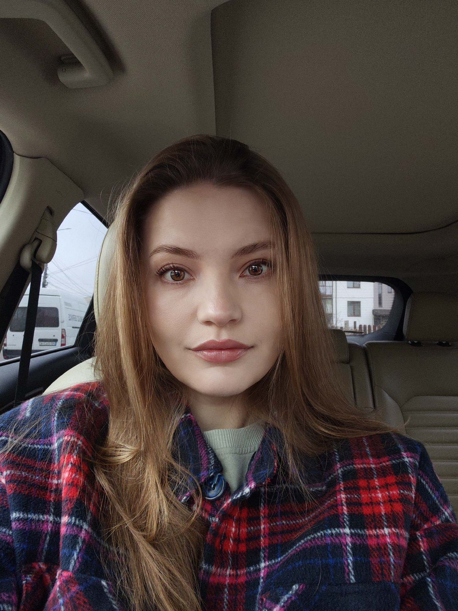 A young woman with auburn hair sits inside a car, presumably in the city of Vinnytsia, Ukraine, as evidenced by a white delivery van visible through the car window, with a license plate number that includes the letters "AT" which are indicative of the Vinnytsia Oblast region of Ukraine. She is wearing a red, blue, and white plaid shirt over a green sweater. The car has beige upholstery. The woman is looking directly at the camera. The view through the car windows reveals a street with buildings.