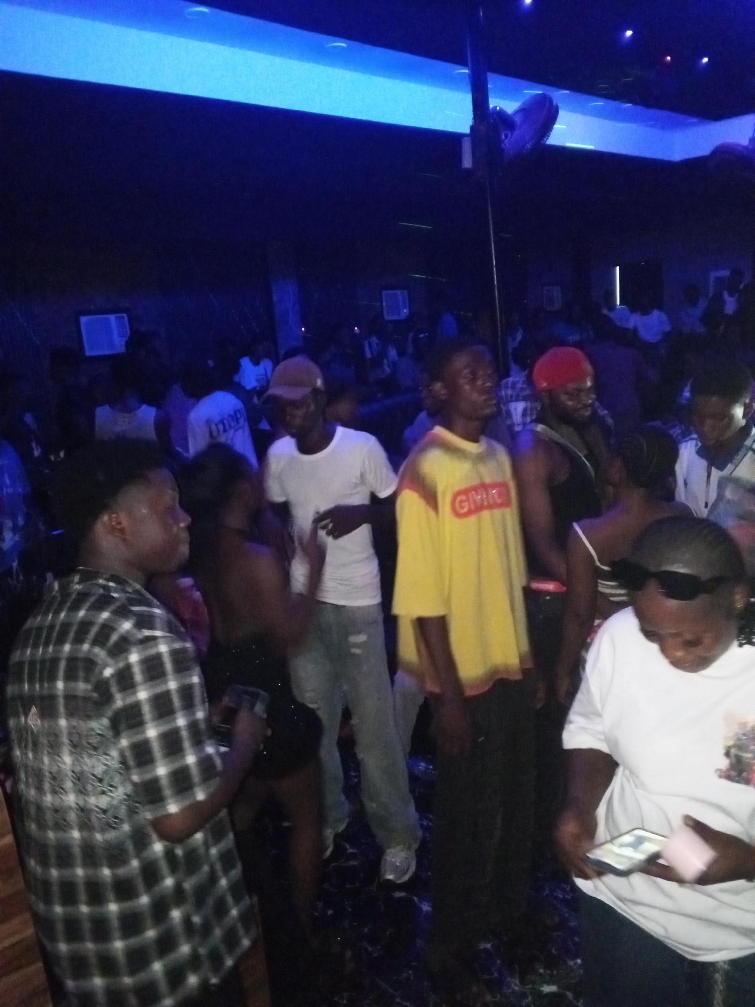 The image shows a crowded indoor scene in Lokoja, Nigeria, with blue lighting. Many people are present, some standing and others interacting. People are wearing various outfits, including t-shirts, plaid shirts, and tank tops. Several individuals are holding phones. A ceiling fan is visible. Air conditioning units are installed on the wall. The overall atmosphere appears to be that of a nightclub or party.