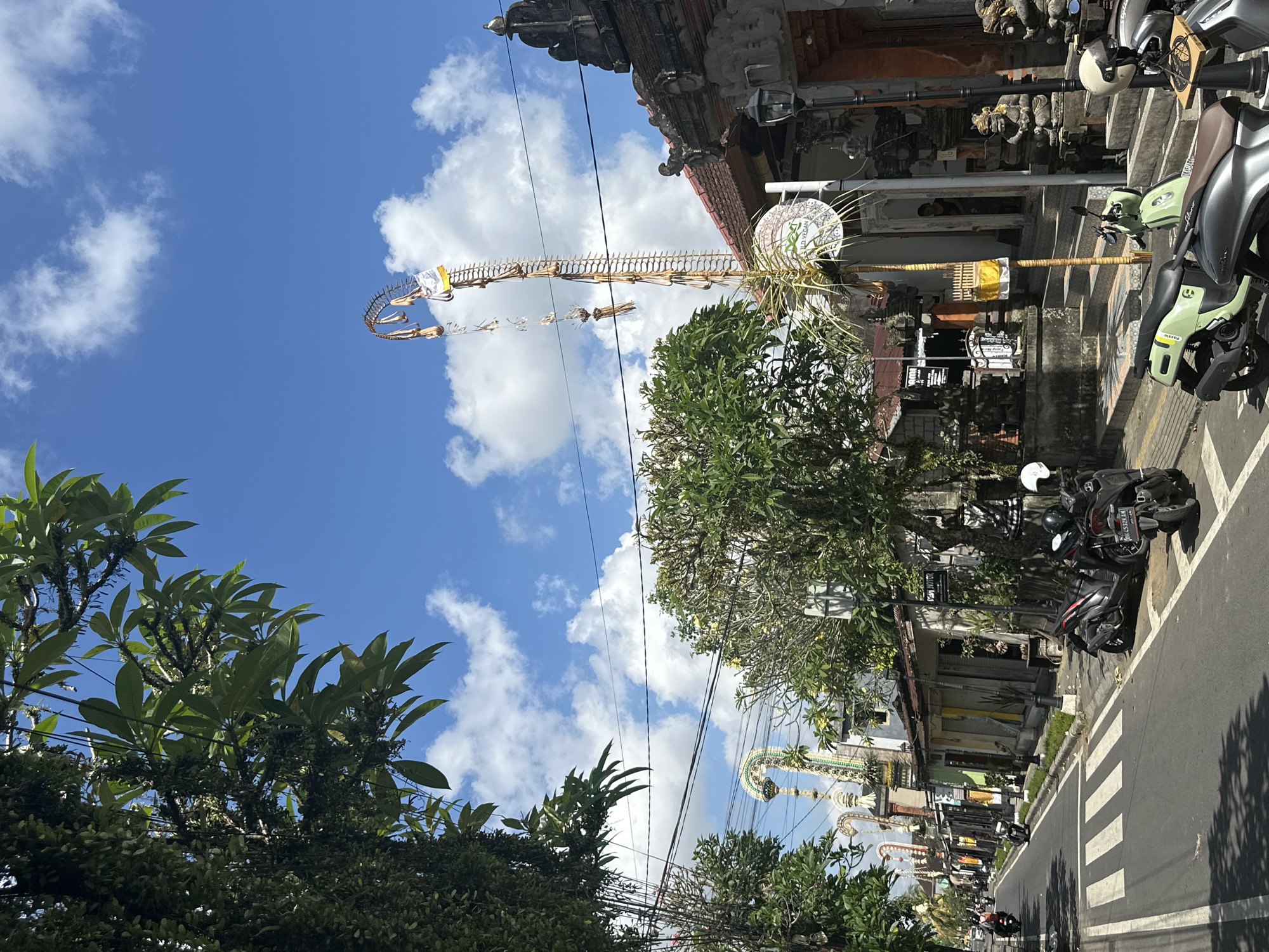 The image depicts a street scene in Ubud District, Indonesia, under a bright blue sky with scattered clouds. Lush green trees line the street, with some hanging over the road, and overhead wires are visible. Several motorbikes are parked along the roadside, and some are being ridden on the street. Traditional Balinese architecture, characterized by ornate stonework and tiled roofs, is evident in the buildings lining the street. A tall, decorative bamboo structure adorned with woven materials and offerings stands prominently in the foreground, characteristic of religious decorations in Bali. People are sparsely visible, with one individual standing near a doorway in the background and others riding motorbikes. The street itself is paved with asphalt and marked with a pedestrian crossing. The overall impression is of a lively street in a tropical urban environment with strong cultural elements.