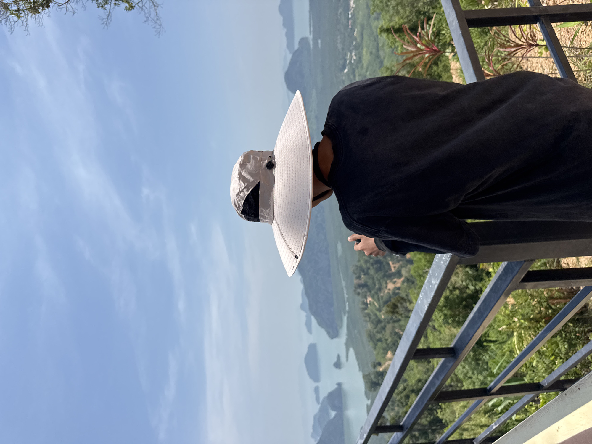 A medium, eye-level shot oriented vertically shows a person wearing a large, light-colored hat and dark clothing, standing at a railing and looking out over a body of water dotted with small, tree-covered islands. The person is positioned on the right side of the frame, leaning forward slightly against the railing. The railing is made of dark metal bars. In the background, there is a blue sky with light clouds to the left, and the islands stretch into the distance with trees visible. The location is likely near Ban Khlong Khian บ้านคลองเคียน, Thailand, given the landscape and vegetation.