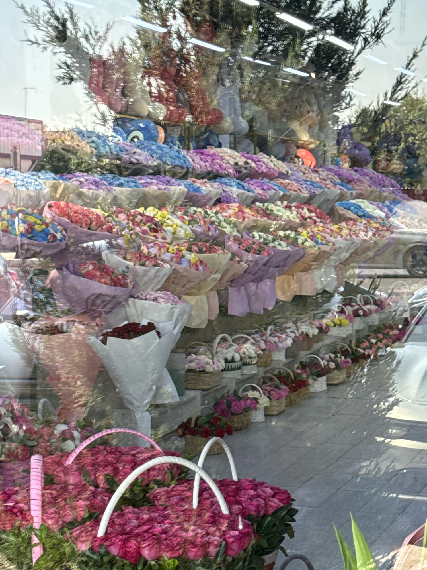 The image shows a large display of flowers, likely in a flower shop or market, in Tashkent, Uzbekistan. The flowers are arranged in various bouquets and baskets, with a focus on roses in shades of pink and red in the foreground. Behind these, there are rows of smaller bouquets in various colors, including pink, purple, blue, yellow, and white. The reflection in the glass storefront shows trees, lighting fixtures, and a blue cartoon character, suggesting the display is inside a shop with a window facing outwards. The overall impression is a vibrant and colorful floral arrangement.