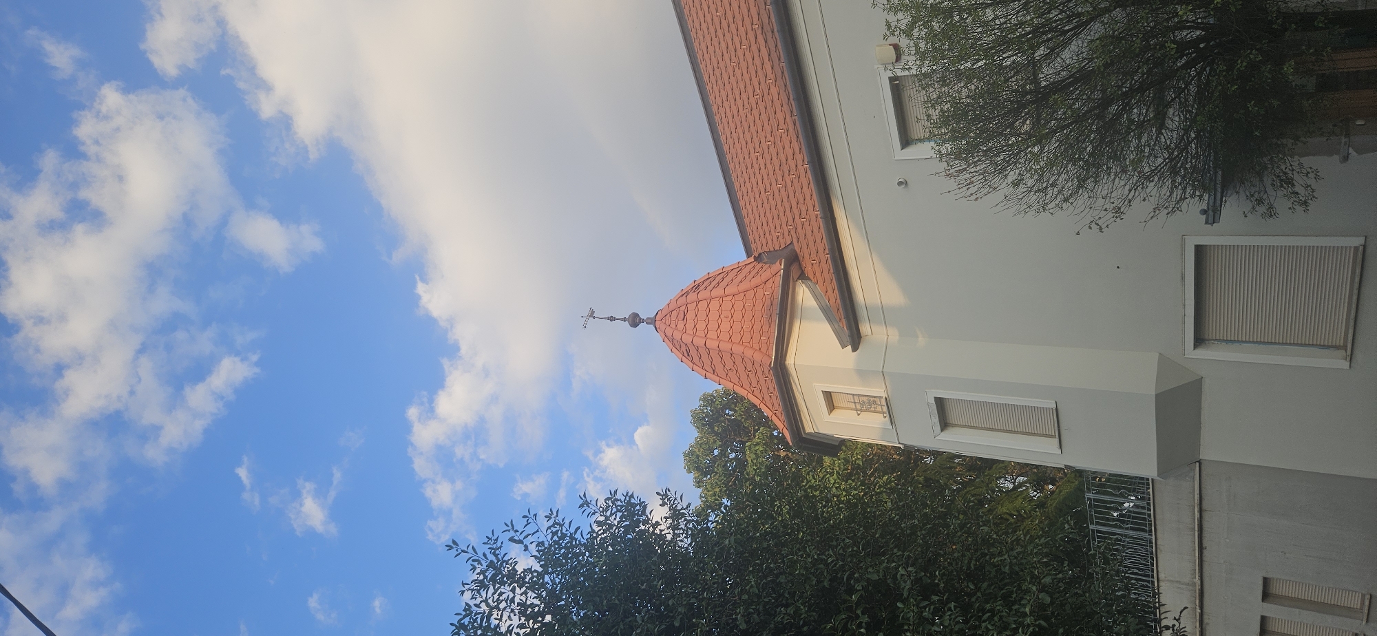 The image depicts a building with a distinctive turret, featuring a red tiled roof and a weather vane. The building is light gray and has several windows, some with closed shutters. Lush green trees surround the building, and a cloudy blue sky is visible above. The scene is bathed in a warm light, suggesting it was taken during either sunrise or sunset.
This building is located in Hévíz, Hungary. Specifically, this is a view of the "Károlyi Apartment House" on Kossuth Lajos utca, a street in the center of the town known for its thermal lake. The architecture suggests it might be an older, possibly historic, residential building or a guesthouse. The presence of trees and the clear sky indicate an outdoor setting in a developed area, likely a residential or tourist zone within the city. No people are visible in the image.