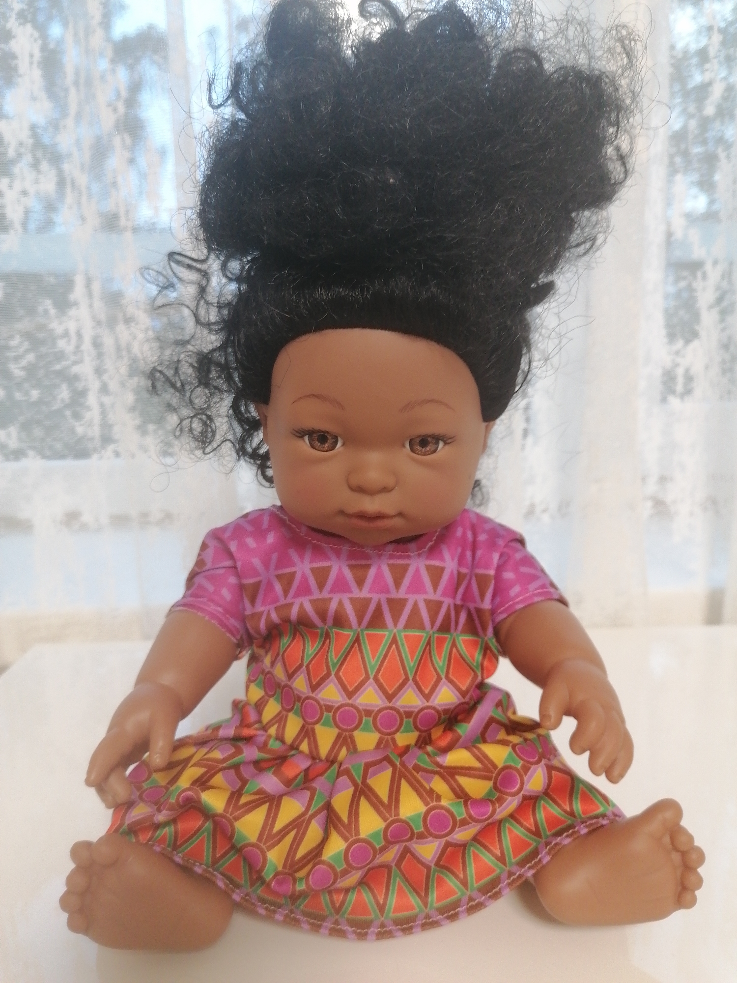 This is a medium shot, eye-level, indoor photograph of a doll. The doll is sitting on a white surface, possibly a table or floor. It has dark, curly hair styled in a high puff. The doll has brown skin, brown eyes, and is wearing a colorful dress with a geometric pattern. The dress is pink with triangular and circular motifs in shades of purple, orange, yellow, and green. Behind the doll, a sheer white curtain with a floral pattern is visible, suggesting a window is present, allowing some natural light to filter through. The background is softly blurred, emphasizing the doll as the main subject. There is no indication of Lagerevo, Russia, in the image.