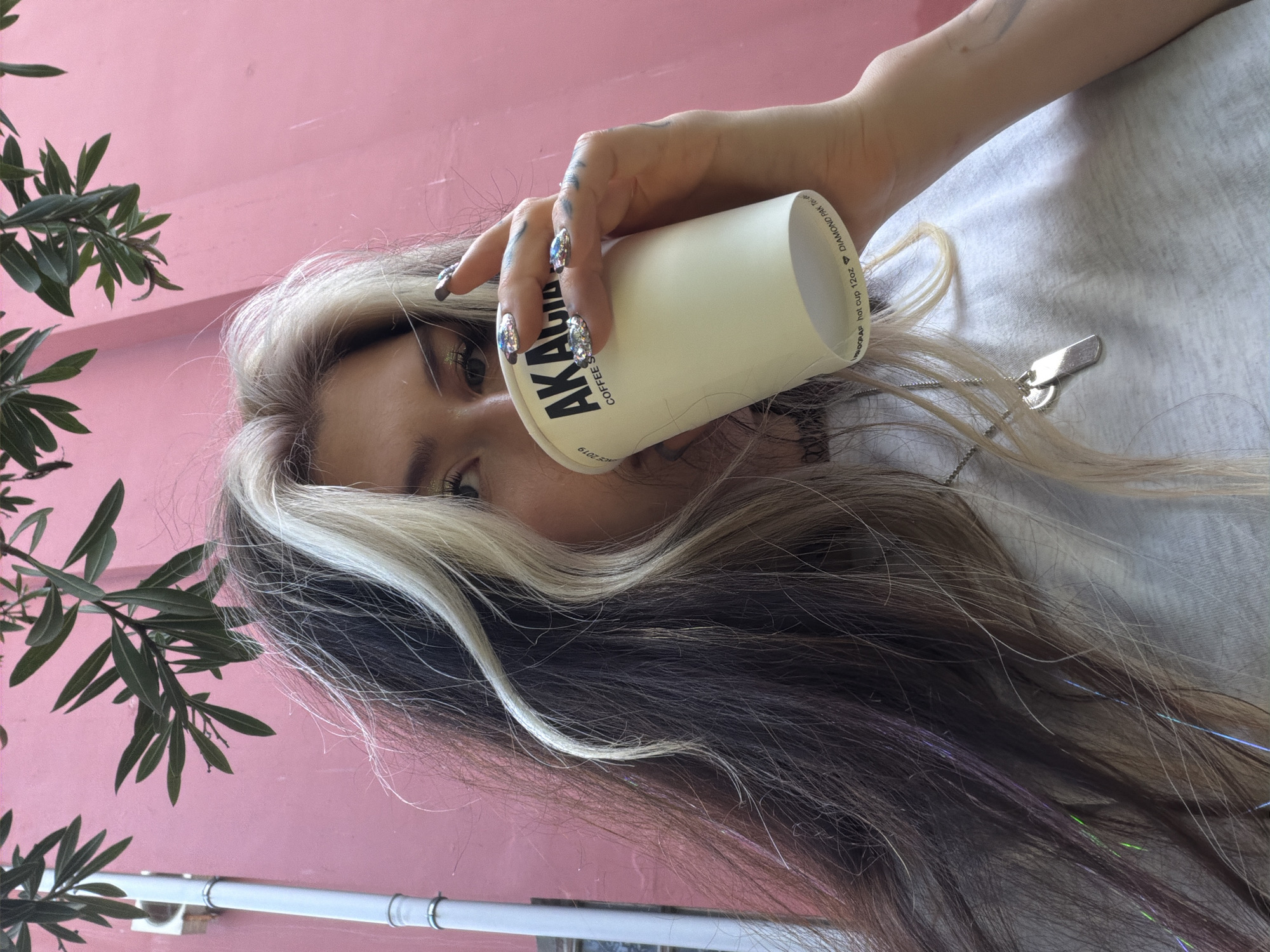 The image, rotated 90 degrees counter-clockwise, features a person holding a white paper coffee cup, partially obscuring their face. One eye is clearly visible, looking directly at the camera, adorned with subtle glittery eye makeup. The person has striking two-toned hair: dark brown or black with a prominent, wide streak of light blonde or platinum hair framing their face. Fine iridescent tinsel strands in purple, green, and blue hues are woven into the darker hair.
The person's hand, holding the cup, showcases elaborately decorated, long fingernails that appear glittery, chrome, or highly reflective. They are wearing a light-colored t-shirt, and a silver chain with a rectangular metallic charm (possibly a key or tag) rests on their chest.
The white coffee cup has black text, partially readable as "AKAR COFFEE S" and "CE 2019". Smaller text on the rim includes "DIAMOND PAK TL" and "12oz HOT CUP TL".
The background consists of a vibrant pink wall. On the left side, green leaves and branches of a plant, possibly an oleander, are visible against the pink backdrop. A white pipe or railing runs horizontally along the lower portion of the image.
The scene depicts a casual moment, likely outdoors in a bright, daytime setting in Budva, Montenegro, perhaps at a cafe or outdoor urban area. The overall impression is stylish and direct, with a focus on personal details and the coffee.