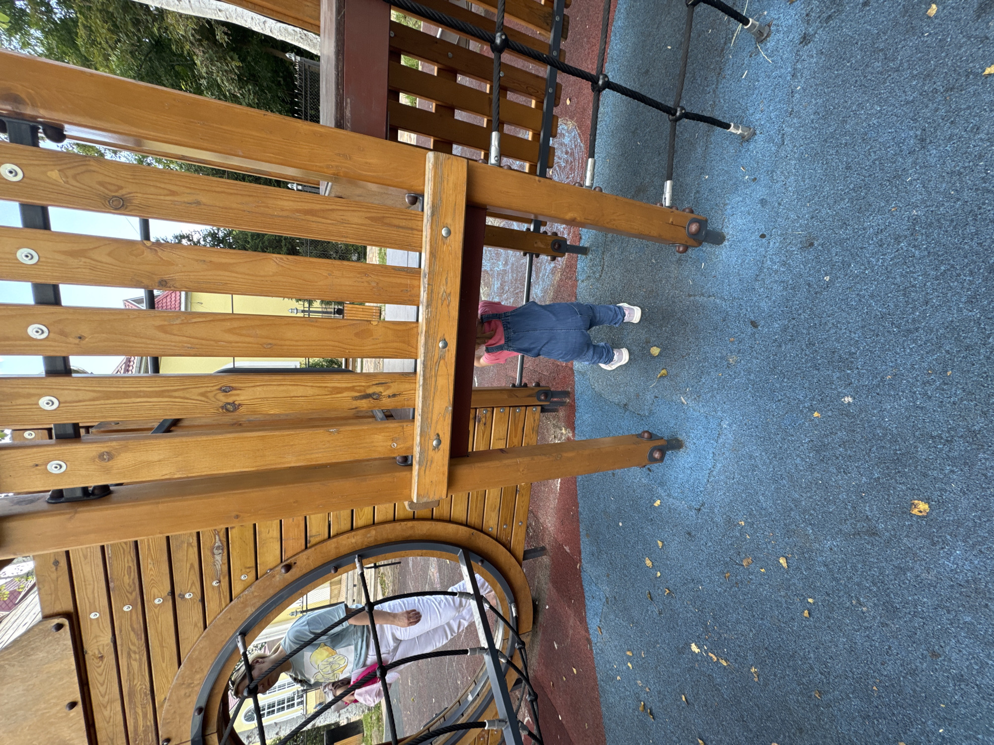 The image shows a children's playground in Mikhaylovsk, Russia. A young child wearing denim overalls and pink sneakers stands beneath a wooden playground structure. The structure is made of natural wood with visible grain and metal fasteners. To the left, a person wearing white pants and a light blue t-shirt with a lemon graphic is partially visible, reflected in a circular wooden frame on the playground. The ground is covered with a blue rubberized surface, scattered with a few fallen leaves. In the background, a yellow building with a red roof and trees are visible, suggesting an outdoor setting within a residential area. The overall scene depicts a typical day at a public playground.