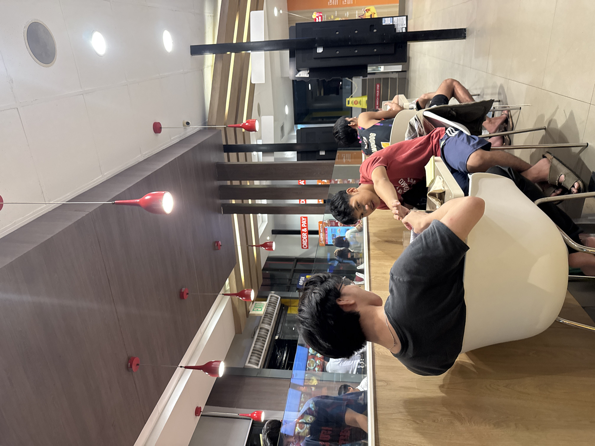 The image captures an indoor scene within what appears to be a modern restaurant or food court. The perspective is from behind a young man with dark hair, wearing a black t-shirt, seated at a light-colored table or counter on a white chair. To his left, another young man in a red t-shirt is visible, looking towards the foreground. Further into the background, other individuals are seated; one wears a black tank top with white lettering, and another's legs are visible, clad in shorts and sandals.
The setting is characterized by light-colored ceilings with multiple recessed lights and several distinctive red, conical pendant lamps hanging at various lengths. Wooden wall paneling runs along one side of the room. There are glass partitions and a serving or ordering counter in the background, where a large black structure or screen is prominent.
Visible text elements include "ORDER & PAY" on a red sign, "Super" as part of a logo, and "EXIT" on a green sign. The phrase "ONE MORE" is discernible on the black tank top of one person. The bright artificial lighting suggests an active commercial environment, possibly during the day or evening. The people appear relaxed, engaged in casual activity, likely dining or waiting for an order.