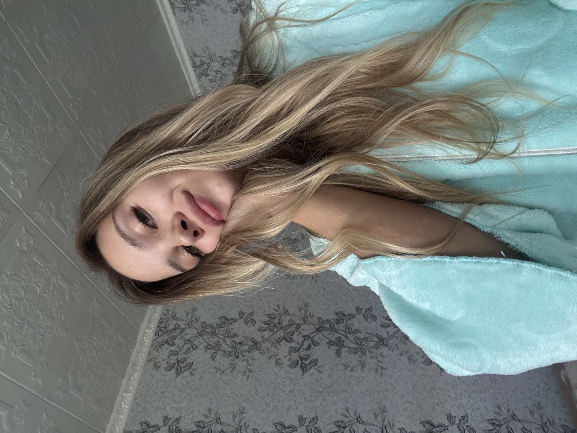 A young Caucasian woman with blonde hair, wearing a light blue robe, is pictured indoors. She is leaning her head on her hand, looking at the camera with her eyes slightly closed and a slight smile. The background consists of a patterned wallpaper and a textured ceiling. The image was taken from a low angle.