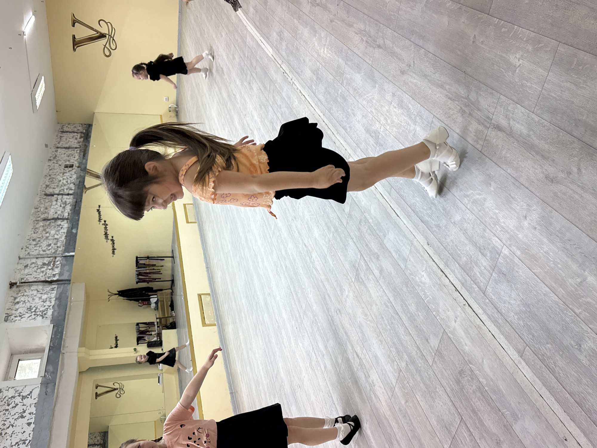 The image captures three young girls in what appears to be a brightly lit dance studio in Chaikovsky, Russia. The room features light grey wooden floorboards, light yellow walls, and large mirrored surfaces that reflect parts of the room, other girls, and a clothes rack.
In the foreground, a girl with long brown hair tied in a ponytail is visible, wearing an orange top with patterned details and a black skirt. She is smiling softly and looking downwards, with her body posed as if in mid-dance or preparing for a movement. Her feet are encased in white dance slippers or similar footwear.
Further back, in the mid-ground, another girl in a black top and skirt is seen in a similar dance-like pose, also wearing white shoes. Her hair is tied up. In the bottom left of the frame, partially visible, a third girl wears a light pink top and black skirt, with one arm raised in a pose.
The setting is clearly a practice space, indicated by the mirrored walls, the type of flooring suitable for dance, and the girls' attire and postures. A distinctive, ornate gold-colored "V" shaped emblem adorns the yellow wall, possibly a studio logo. Rectangular light fixtures are visible on the ceiling, and some sections of the walls have a rough, textured appearance, possibly exposed brick or concrete. The overall atmosphere suggests a cheerful and focused dance lesson or rehearsal during the daytime.