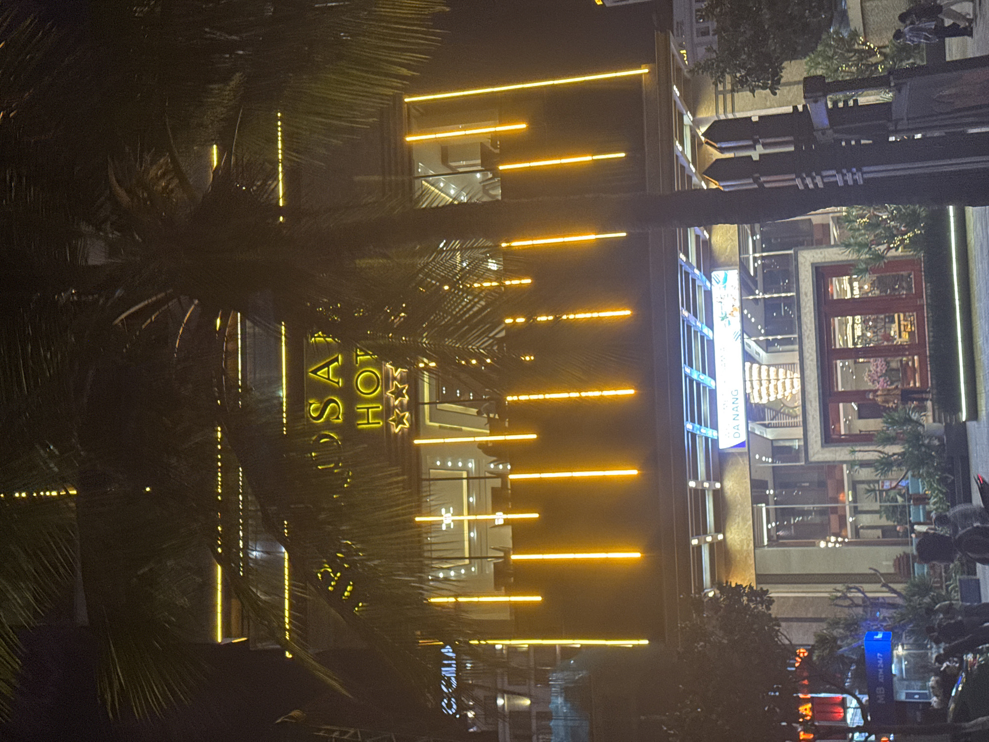 The image depicts a nighttime view of the Adamo Hotel in Đà Nẵng, Vietnam. The hotel's facade is illuminated with linear lights, creating a distinct visual pattern. A palm tree is visible in the foreground, partially obscuring the view of the hotel. Pedestrians and vehicles can be seen in the street in front of the hotel. The surrounding buildings are also illuminated, contributing to the urban environment.