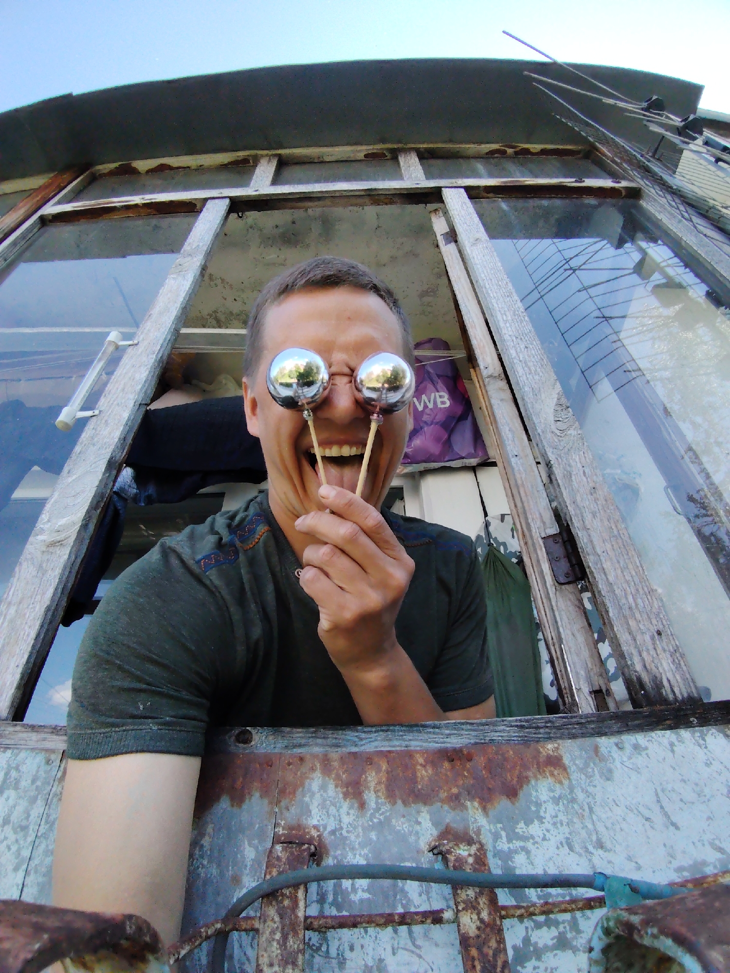 A low-angle, wide-shot captures a person sticking out of a weathered window frame, holding two silver spheres up to their eyes. The person, with short, dark hair and an open mouth with their tongue sticking out, wears a dark green t-shirt with a patterned neckline. The window frame is constructed of rough, aged wood, with visible rust and peeling paint on the surrounding metal structure. The background shows a clear sky and what appears to be a rural or semi-abandoned setting. The specific location in Chaikovsky, Russia, is not identifiable from the visual elements present.