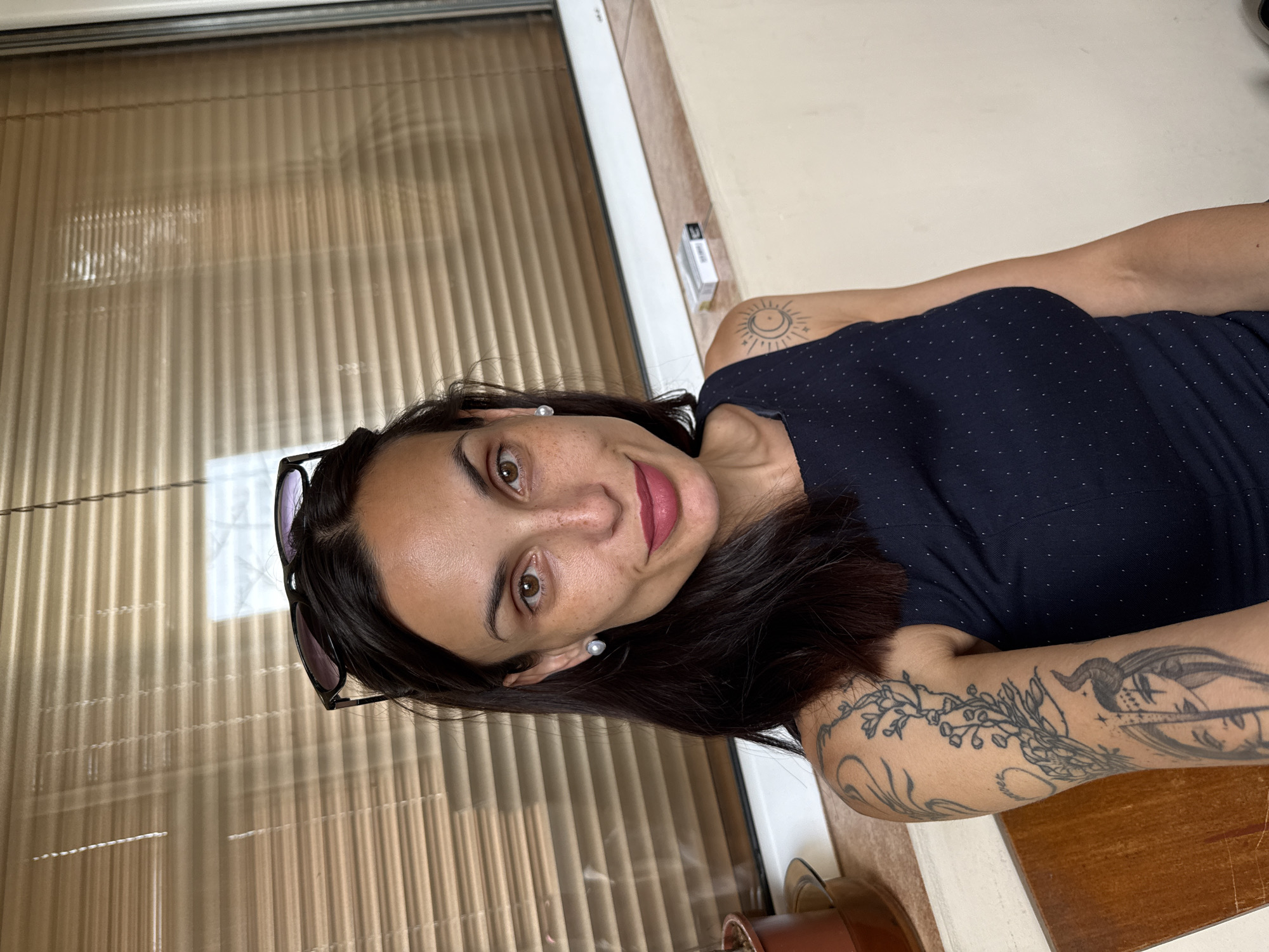 A woman is posing for a selfie. She has dark hair, brown eyes, and fair skin. She is wearing a navy blue top with small white dots. Sunglasses are perched on her head. Tattoos are visible on her arms and shoulder. She is smiling slightly, wearing lipstick. Behind her is a window with beige blinds. A small white box is visible on the window sill. Based on the presence of writing in Bulgarian, and a previous query, it is highly likely that this image was taken in Pleven, Bulgaria.