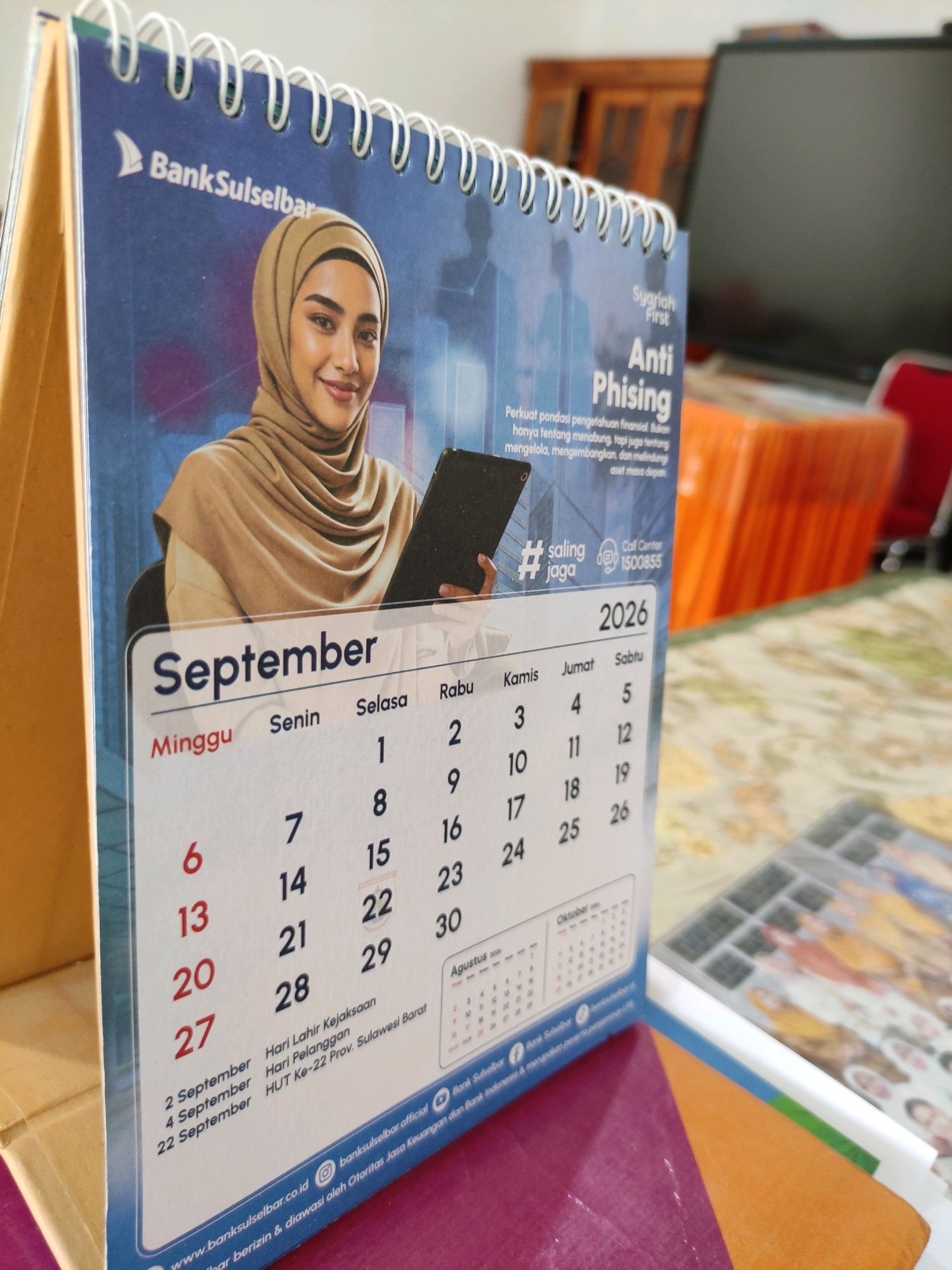 This is a close-up, eye-level shot of a desk calendar for September 2026. The calendar features a photograph of a woman wearing a hijab. The calendar is open to a page displaying the month of September, with dates numbered from 1 to 30. The days of the week, Sunday through Saturday, are listed at the top of the calendar grid. The calendar also includes important dates such as "Hari Lahir Kejaksaan" (Prosecution Birthday) on September 2nd, "Hari Pelanggan" (Customer Day) on September 4th, and "HUT Ke-22 Prov. Sulawesi Barat" (22nd Anniversary of West Sulawesi Province) on September 22nd. The bank's logo, "BankSulselbar," is visible at the top of the calendar. The background is blurred, showing a desk with some papers and a computer monitor. The scene is indoors. The image's composition and the presence of the "HUT Ke-22 Prov. Sulawesi Barat" marking strongly suggest the location is in Bulukumba, Sulawesi Selatan, Indonesia, as the province's anniversary would be relevant to local institutions and events.