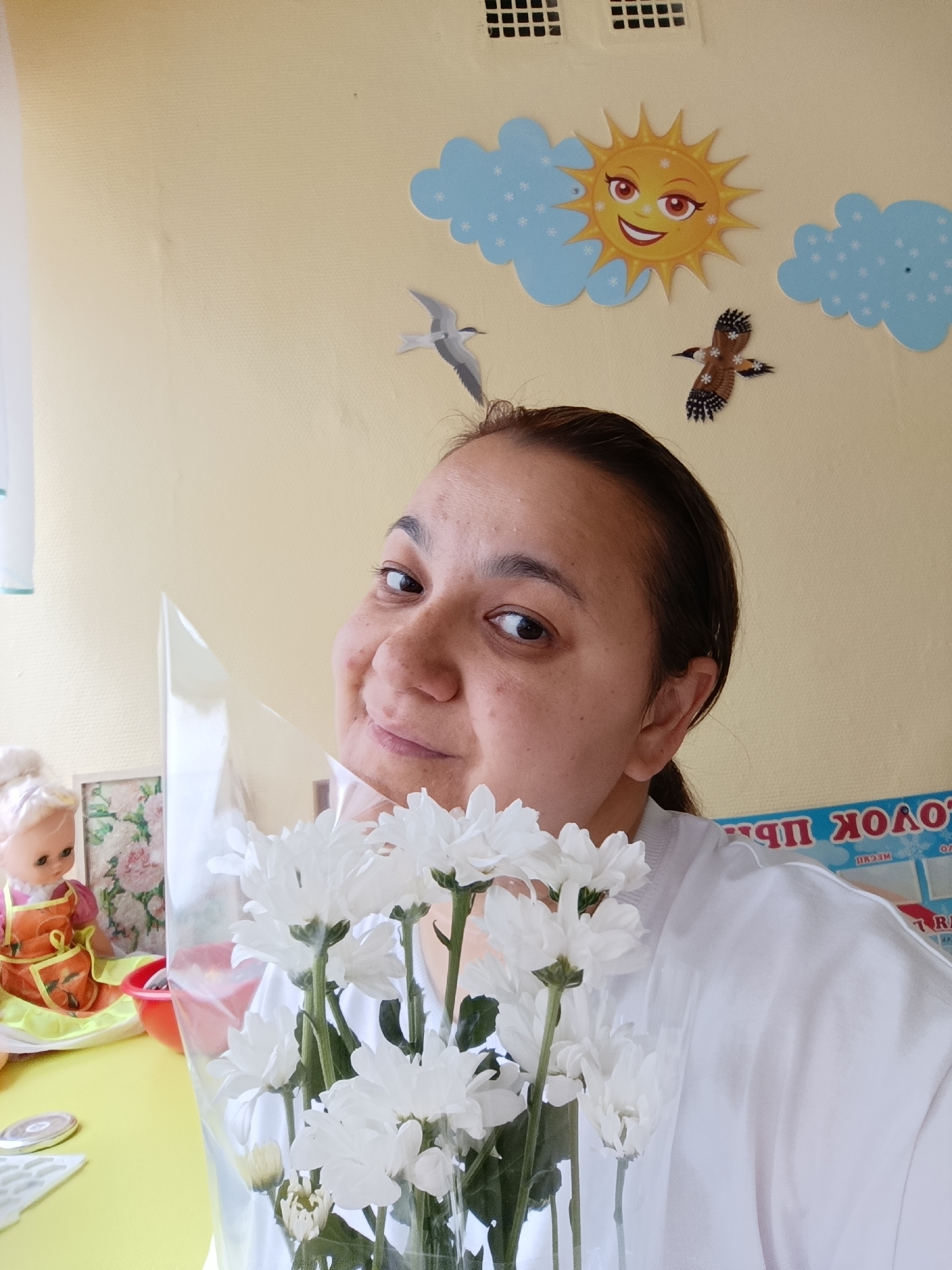 The photograph shows a woman holding a bouquet of white chrysanthemums. The background consists of a yellow wall decorated with cartoon-like images of a sun with a smiling face, blue clouds with snowflake designs, and paper cutouts of birds. In the periphery, a doll is visible on a table. The location is Chaikovsky, Russia.