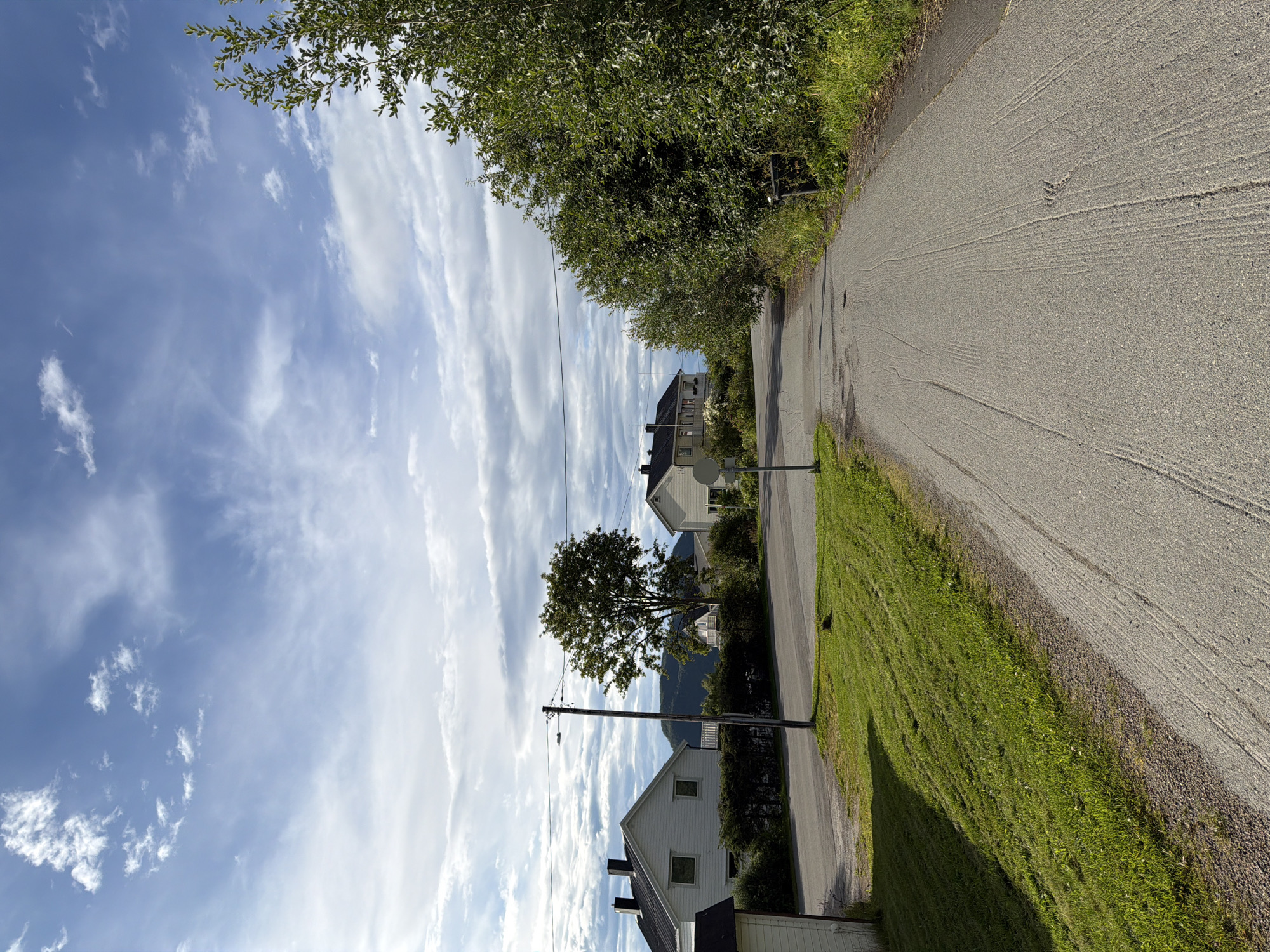 The image depicts a rural road in Harstad, Norway, on a cloudy day. The road is paved and appears to be a residential street, with houses visible on either side. The houses are white with dark roofs. Green grass covers the sides of the road, and trees line the right side of the image. In the distance, a mountain can be seen through the trees. There are no people visible in the image. The sky is a light blue with white clouds.