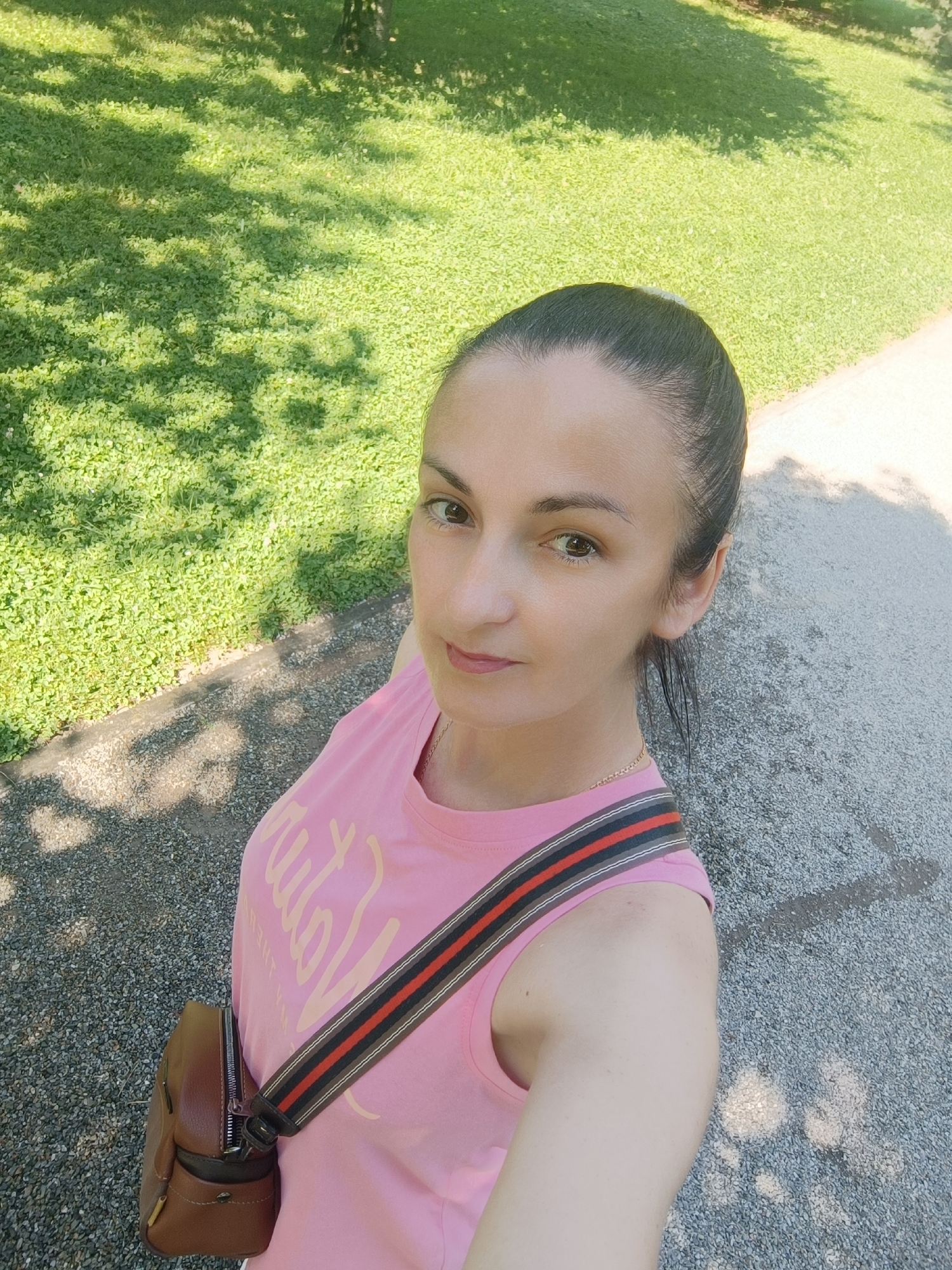 A medium shot from above shows a woman in a pink sleeveless t-shirt and a brown crossbody bag in a park. She has her dark hair pulled back into a ponytail, and she is looking up and to her right. Her t-shirt has an unreadable graphic in a light pink color, and a colorful strap with red, black, and gray stripes goes across her chest. The background is a grassy area dappled with sunlight and shadows, with a paved path running along the right side of the frame. The scene appears to be a selfie taken outdoors in a public space. This image was taken in Târgu Jiu, Romania.