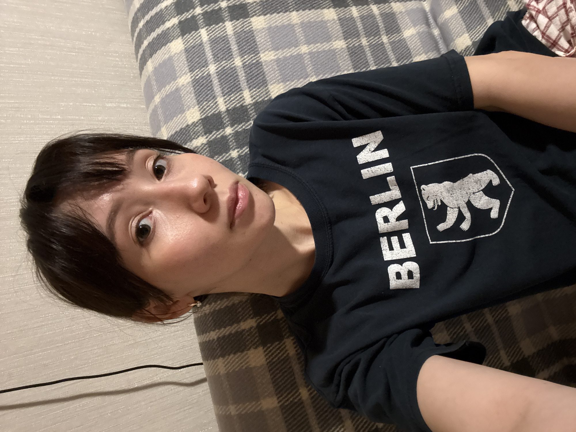 A woman with dark brown hair and fair skin is lying on a patterned, gray and white plaid surface. She is wearing a dark blue t-shirt with "BERLIN" printed in white letters and a white bear emblem. A black cable runs across the lower left of the frame. The location is indeterminate and there is no indication it is in Kazan, Russia.