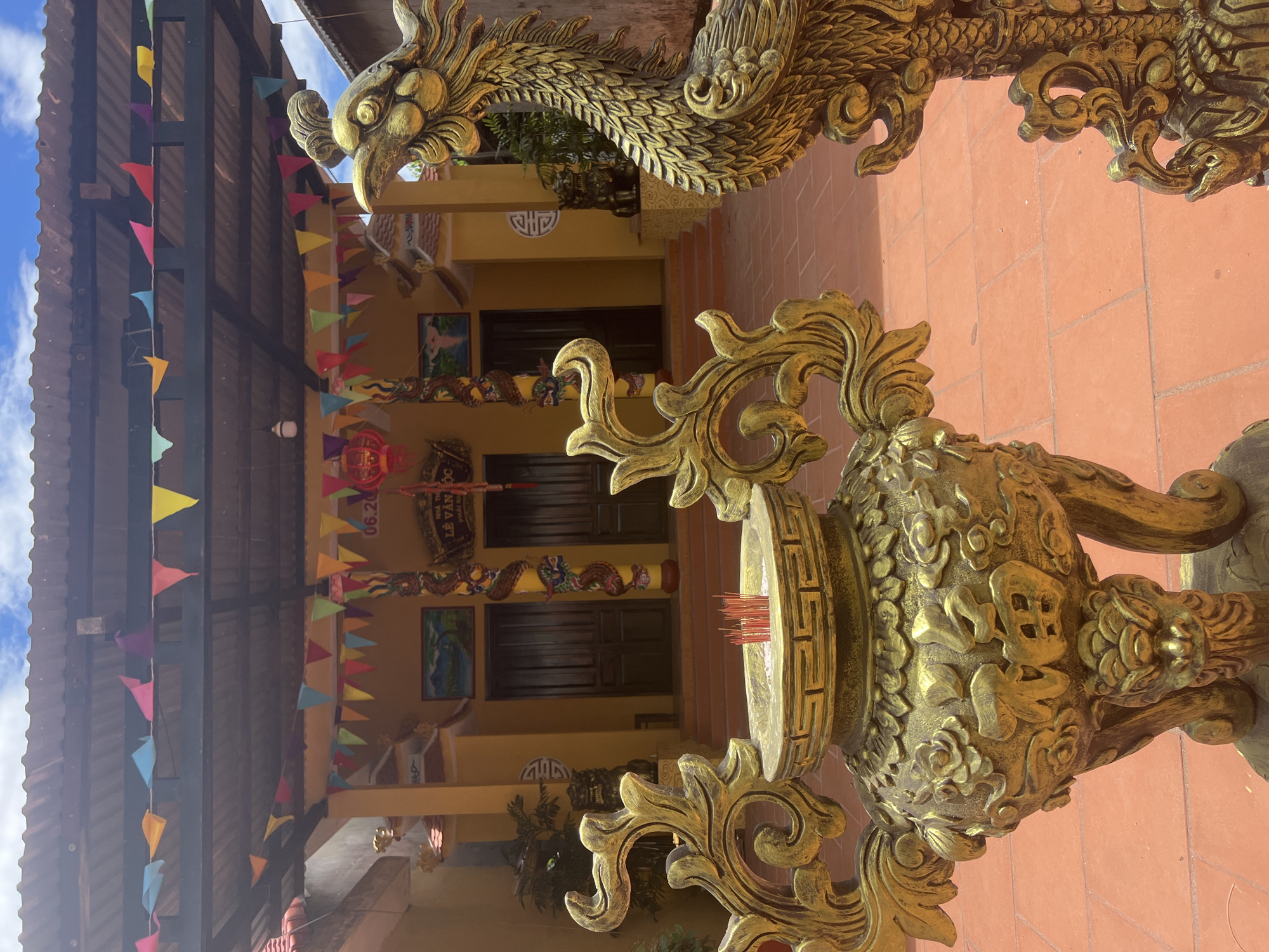 This image captures the exterior of a Buddhist temple in Hoi An, Vietnam. In the foreground, two ornate golden statues are prominently featured: a large incense burner filled with red incense sticks on the left, and a detailed phoenix or dragon statue on the right. The temple itself, painted a vibrant yellow, stands in the background with two large wooden doors leading inside. Colorful triangular flags are strung across the roofline and above the entrance, suggesting a festive occasion. The courtyard is paved with red tiles. The sky is visible above, with some clouds. The architectural style and decorations are characteristic of Vietnamese temples.