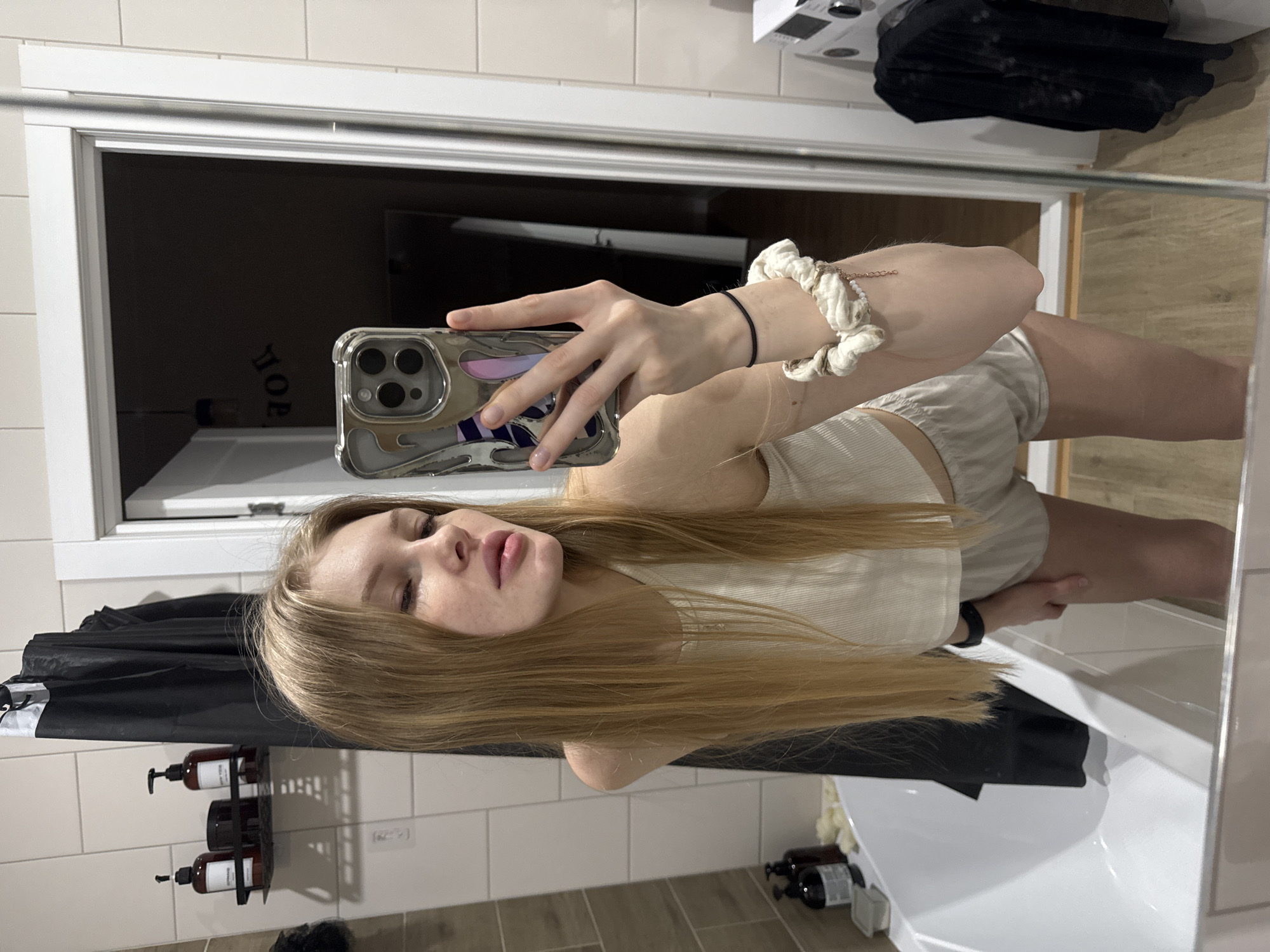 A young woman is taking a selfie in a bathroom. She is holding a smartphone in her left hand, with a white scrunchie on her wrist. She has long blonde hair and is wearing a light-colored top and shorts. The bathroom has beige tiled walls and a white bathtub. A black shower curtain is visible, along with a shower caddy holding shampoo bottles. The reflection shows a washing machine in the background. There is no information to determine that the location is in the city of Perm, Russia.