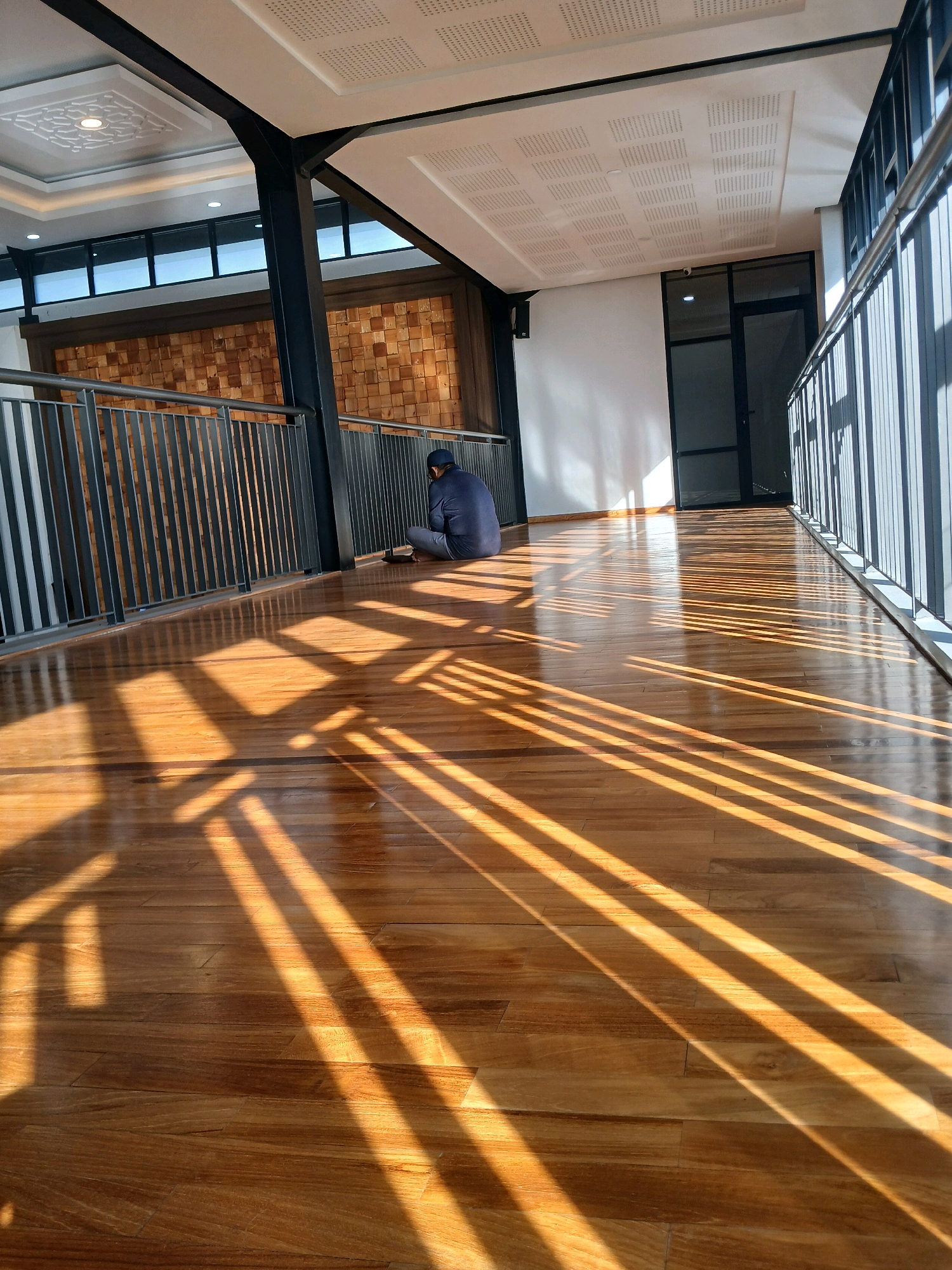 A lone individual, wearing a navy blue long-sleeved shirt and gray pants, is seated on a polished wooden floor, facing away from the camera. They are positioned near a metal railing, with their hands resting on the floor in front of them. The setting appears to be an indoor walkway or hallway, likely part of a modern building in Arjasari, Indonesia. Sunlight streams in through a series of vertical elements, casting long, parallel shadows across the gleaming wooden floor. The walls of the walkway are a neutral white, with a section of textured wooden panels visible in the background. The ceiling is white with a patterned acoustic tile design and recessed lighting. A glass-doored entrance is visible further down the hallway on the right.