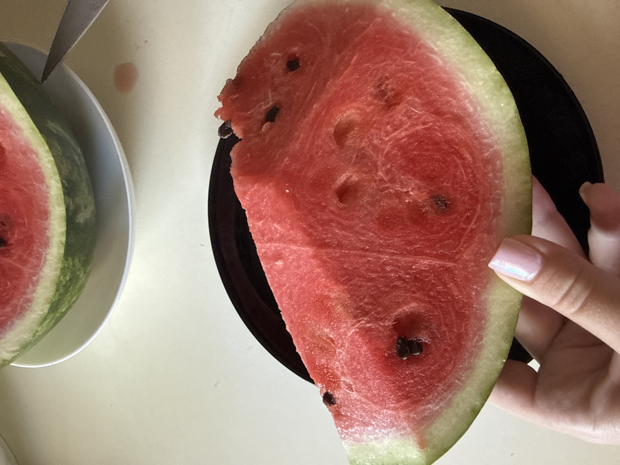 A slice of watermelon is held by a hand with a pale pink manicure against a black plate. The watermelon is a vibrant red with visible seeds and a pale green rind. The background is a light-colored, smooth surface, likely a table, with a sliver of a larger watermelon on a white plate in the upper left corner. A knife is partially visible to the right of the larger watermelon slice, and a small pink liquid spill is present on the table. The scene appears to be indoors, possibly a kitchen or dining area.
