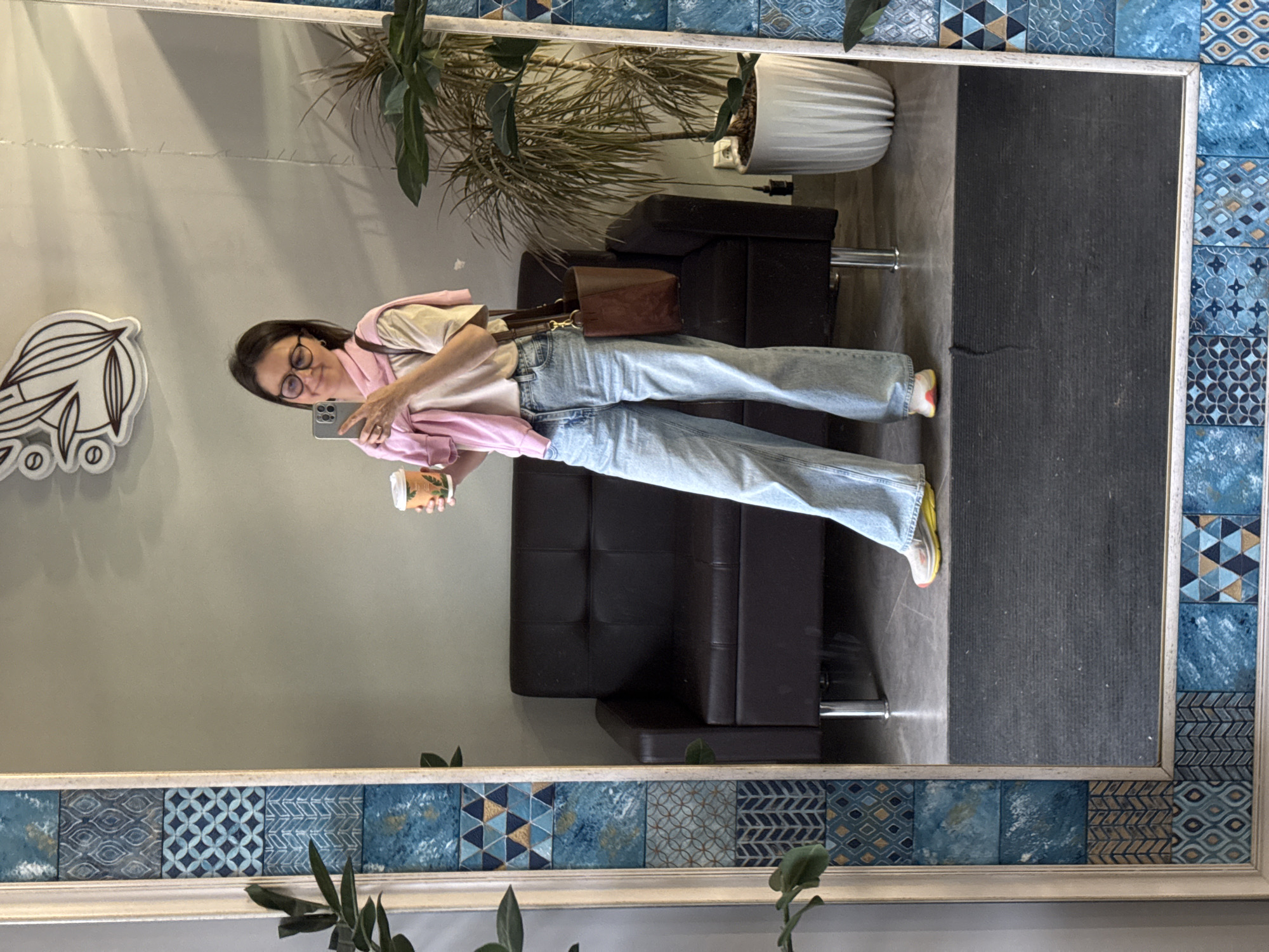 A mirrored reflection shows a woman standing and holding a coffee cup and a smartphone in a cafe in Izhevsk, Russia. She wears glasses, a beige top, a pink sweater draped over her shoulders, light blue jeans, and yellow sneakers. A brown purse hangs from her shoulder. A sign with a stylized plant design is visible on the left. Behind her is a black leather couch and a potted plant in a white pot. The floor is grey, and the mirror is framed with patterned blue tiles.