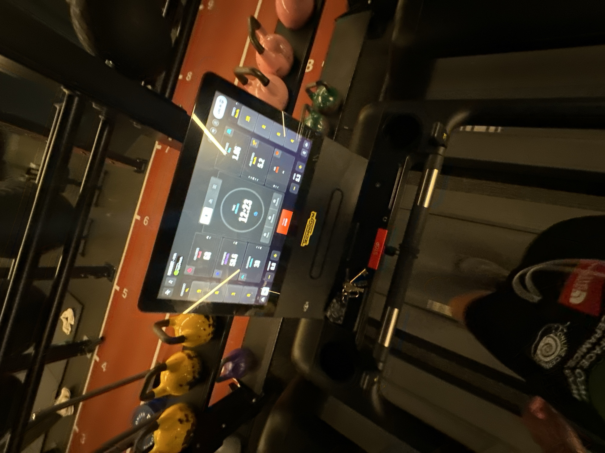 Here is a description of the image:
**Scene:**
The image appears to be taken at a gym or fitness center. The viewpoint is from a slightly elevated, oblique angle, likely near the side of a treadmill.
**Objects:**
* **Treadmill:** A portion of the treadmill is visible, including its console. The console displays digital information, including time ("12:23"), speed ("2.6"), and an elliptical training program interface.
* **Kettlebells:** Several kettlebells of varying colors (pink, green, yellow, blue, purple) are arranged on a rack along the left side. The rack is numbered.
* **Weight rack:** A weight rack is visible to the left of the treadmill.
* **Clothing:** A person is on the treadmill, partially visible in the corner of the image. They are wearing a black baseball cap, the logo is partially obscured.
**Location:**
While the equipment suggests a gym setting, the photograph does not provide any specific indicators of a location in London, UK, or elsewhere. Without additional context, the exact location cannot be determined.