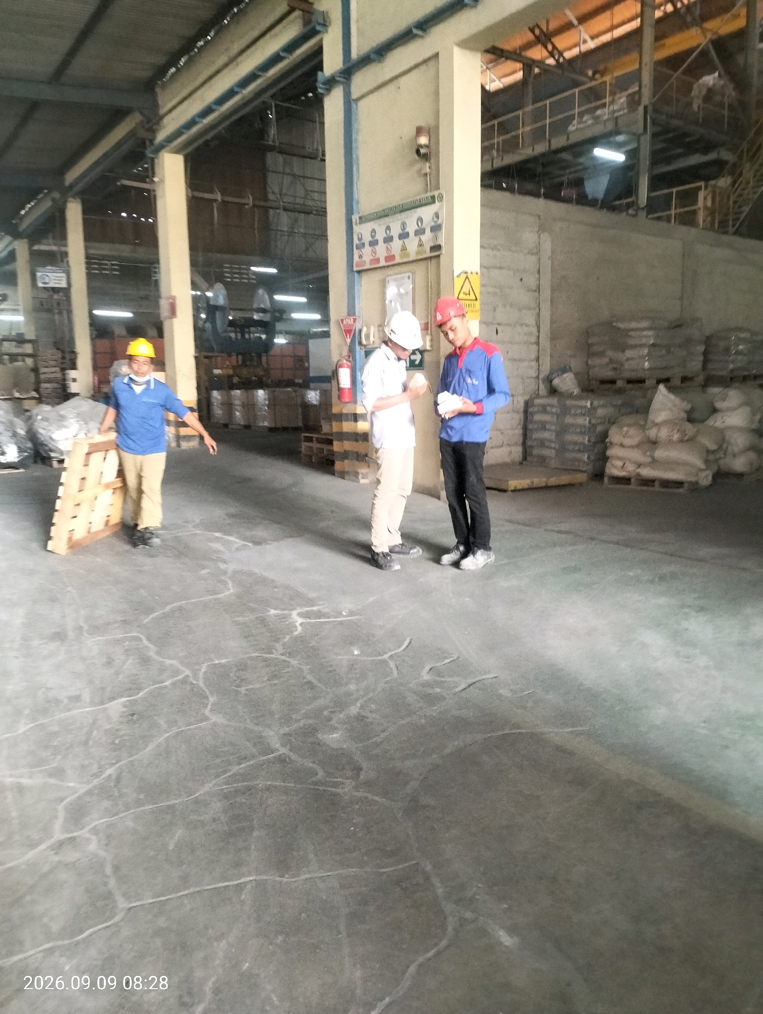 The image shows a warehouse interior with concrete flooring that has a cracked pattern. Three men are present. One man on the left, wearing a yellow hard hat, blue shirt, and tan pants, is holding a wooden pallet. Another man in the center, wearing a white hard hat and white shirt with tan pants, appears to be talking to the man on the right. The man on the right wears a red and blue uniform with a red hard hat and is holding something in his hands. In the background, there are stacks of bagged materials on pallets, and industrial shelving and equipment. A fire extinguisher is visible on the right side of the image. The timestamp on the bottom left corner indicates the image was taken on September 9, 2026, at 08:28. The location is identified as Bambe, Indonesia.