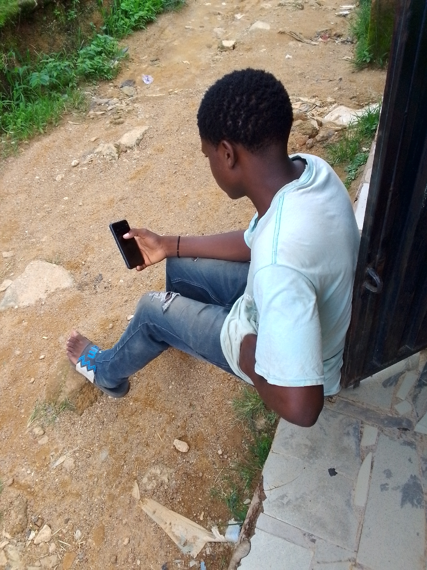 A young Black person is sitting on a stoop, holding a black smartphone. The person is wearing ripped blue jeans and a light blue t-shirt. The background is a dirt path with some grass and debris. The person is looking down at the phone, seemingly engrossed in its use.