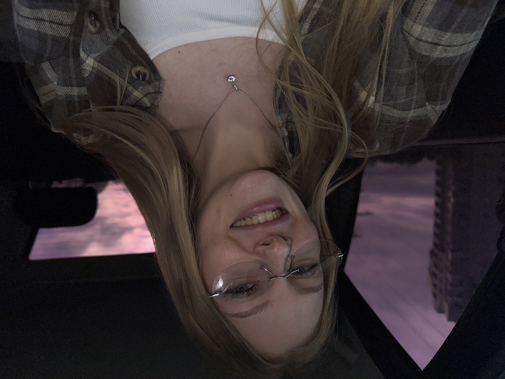 The image shows a person, likely female, with blonde hair wearing rimless glasses and a white tank top under a plaid shirt. A necklace with a small pendant is visible. The person is smiling. The scene is set inside a car at night, with a reflection of buildings and a pinkish sky visible in the car window. Based on the reflections, the location is likely in an urban setting. It cannot be determined with certainty that the image was taken in Perm, Russia based solely on the information available in the image.
