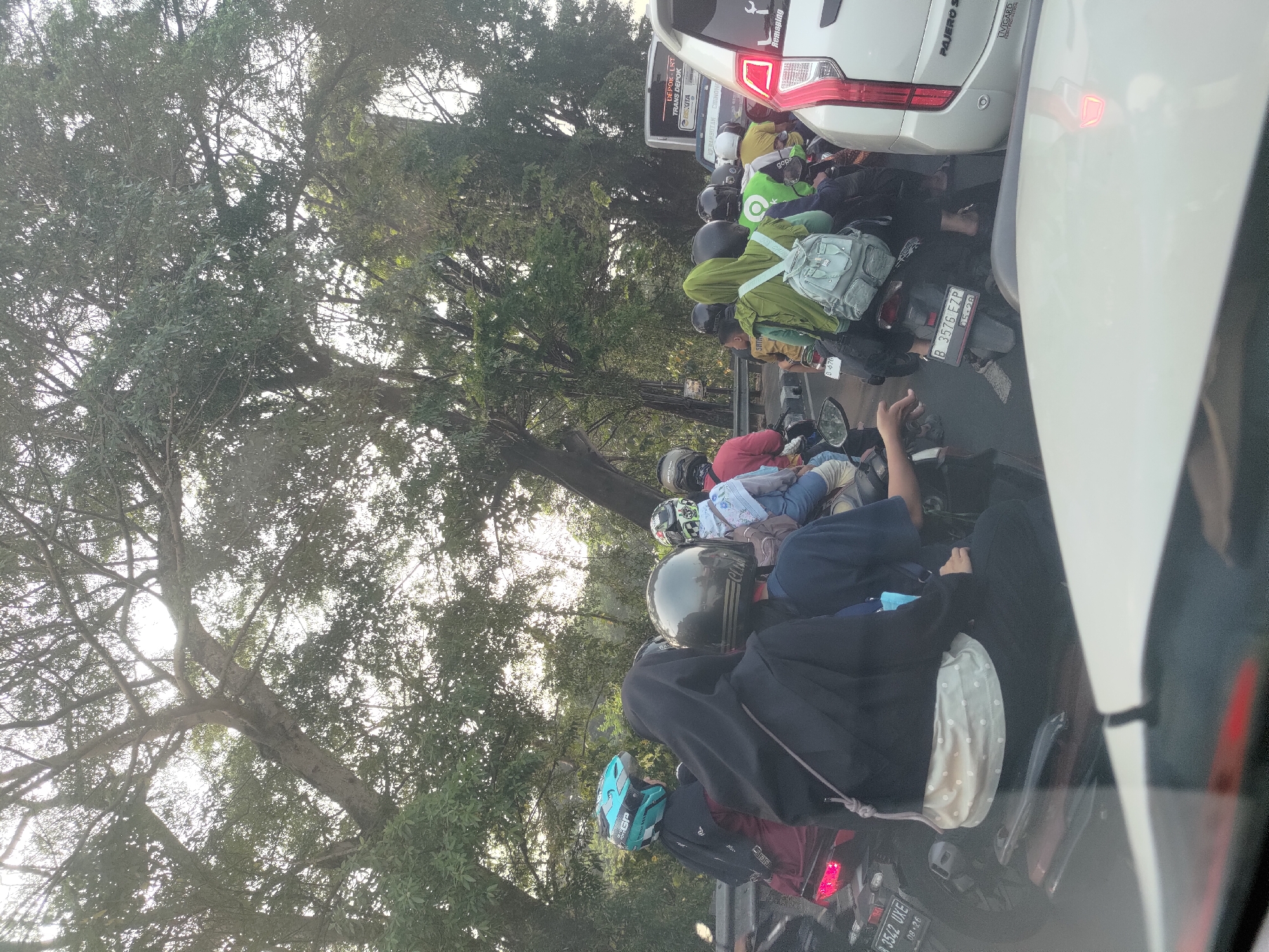 The image shows a traffic jam on a street in Depok, Indonesia. Multiple motorcycles, cars, and a bus are visible. The environment is characterized by lush green trees bordering the road, suggesting a semi-urban or suburban setting. The time of day appears to be late afternoon, with the sun filtering through the tree canopy.
In the foreground, a white car's hood and windshield are visible, indicating the photo is taken from inside a vehicle. Behind this, a line of motorcycles fills the road. Several individuals on these motorcycles are wearing helmets, with some having passengers. Notable details include a motorcycle with a blue helmet, another with a black helmet, and a person wearing a green hijab with a backpack. The license plates of some motorcycles are partially visible. Further back, a white bus with "DEPOT - LRT TRANS DEPOK" written on it is stuck in traffic. The overall scene depicts a common urban traffic congestion scenario in Indonesia.