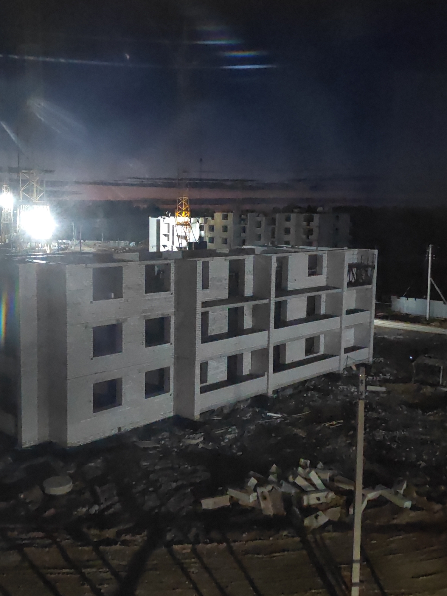 The image depicts a construction site in the city of Cherepovets, Russia, at night. An unfinished multi-story building constructed of white brick stands prominently in the foreground. The building's window spaces are open and unfinished. Scattered around the base of the building are piles of construction debris, including bricks. In the background, other buildings in various stages of construction are visible, illuminated by bright construction lights. Yellow construction cranes are visible amidst the buildings. The sky is dark, with some faint light and atmospheric effects. A pole is on the right side of the image.