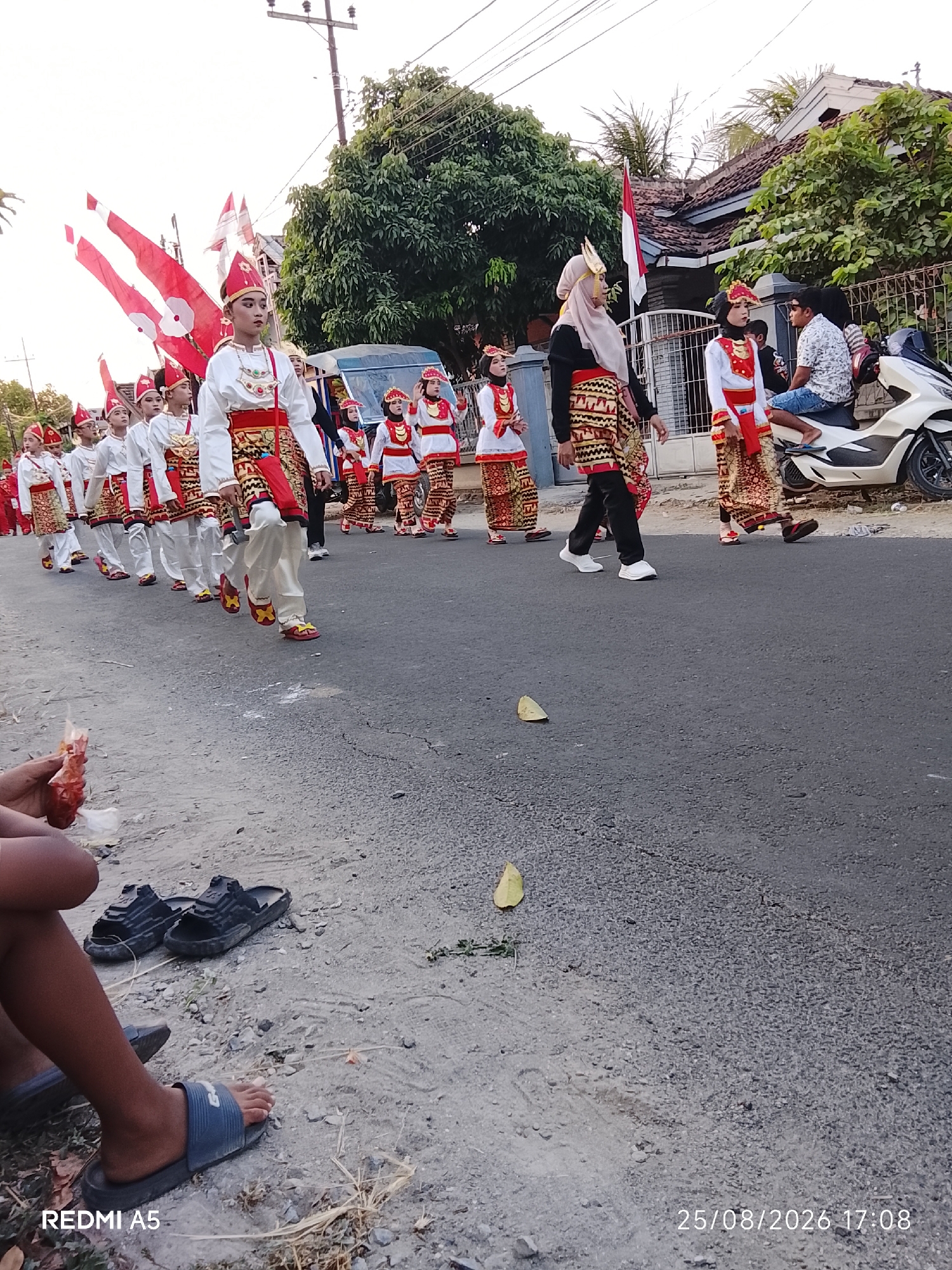 In Semanding, Indonesia, a group of individuals in traditional attire is participating in a parade. The participants, predominantly young people, are adorned in white shirts, red and gold patterned skirts or trousers, and red headwear. Some individuals are carrying large red and white flags. They are walking in an organized procession on a paved road, with buildings and trees lining the street. A white motorcycle is parked on the side of the road, and a few adults are observing the parade. In the foreground, a person's legs and feet are visible, wearing sandals, with a red item held in their hand. The scene appears to be taking place during daylight hours.