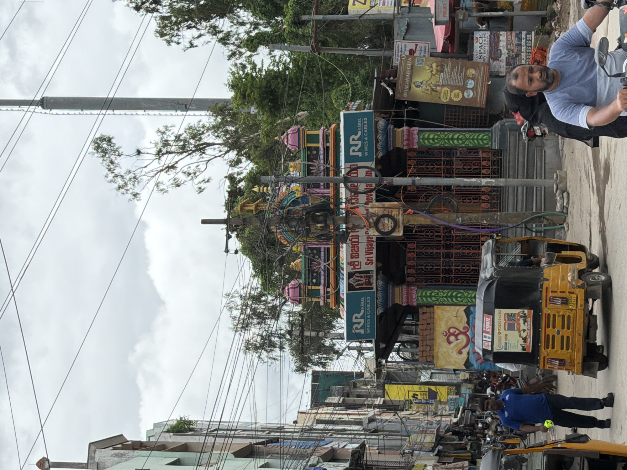 The image captures a street scene in Shamshabad, India, under an overcast sky. The foreground features a bright yellow auto-rickshaw, facing right, with a man in a blue t-shirt walking beside it to the left, holding a yellow object. Behind the auto-rickshaw, a man wearing a light blue t-shirt and a beard rides a motorcycle, looking forward. To the right of the auto-rickshaw, a woman in a black hijab is partially visible.
Dominating the center of the image is a colorful, ornate structure, likely a Hindu temple or shrine, with vibrant pillars, intricate carvings, and tiered rooftops. A sign above the entrance reads "RR KABEL WIRES & CABLES" in English, and also in a regional script. Another sign above that displays "శ్రీ విజయ" and "Sri Vijaya". To the left of this structure, a large yellow sign with Telugu script and an "Om" symbol is visible on the wall. Further to the left, buildings with multiple floors and balconies line the street, with numerous electrical wires crisscrossing the scene. Several other vehicles, including motorcycles and auto-rickshaws, are present further down the street, suggesting a bustling urban environment. Advertisements and signs are plastered on various surfaces. The overall impression is of a busy street in an Indian city with a strong religious presence.