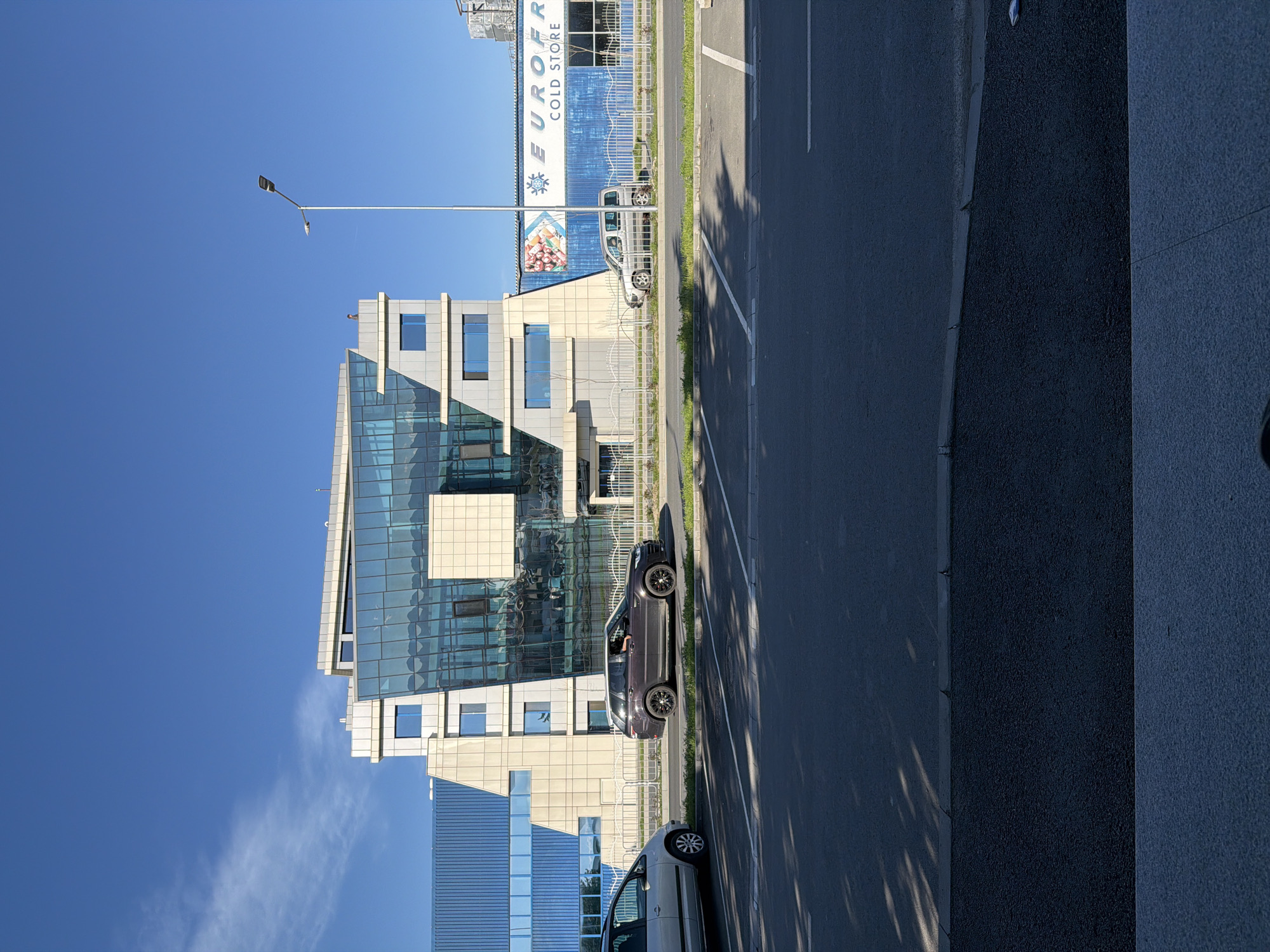 The image shows a building with a modern glass and concrete facade, located on a street in Plovdiv, Bulgaria. A gray car is parked on the side of the road next to the building. Another white car is seen in the front. There is a large industrial building behind it named EUROFRIG COLD STORE. A street lamp is also visible. The sky is clear blue.