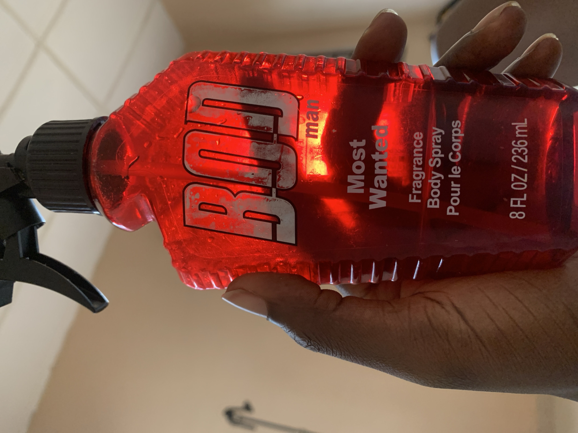A person's hand holds a bright red spray bottle of "BOD man Most Wanted" fragrance body spray. The bottle is translucent, allowing the red liquid inside to be visible. The label on the bottle features the brand name "BOD" in large, stylized silver letters. Below that, smaller text reads "man," "Most Wanted," "Fragrance," "Body Spray," and "Pour le Corps." The bottle also indicates a volume of "8 FL OZ / 236 mL." The person's hand, with dark skin and manicured nails, is gripping the bottle, with fingers partially obscuring the label. The background appears to be a wall with some tiling, suggesting an indoor setting, possibly a bathroom or dressing area. The lighting suggests it is daytime.