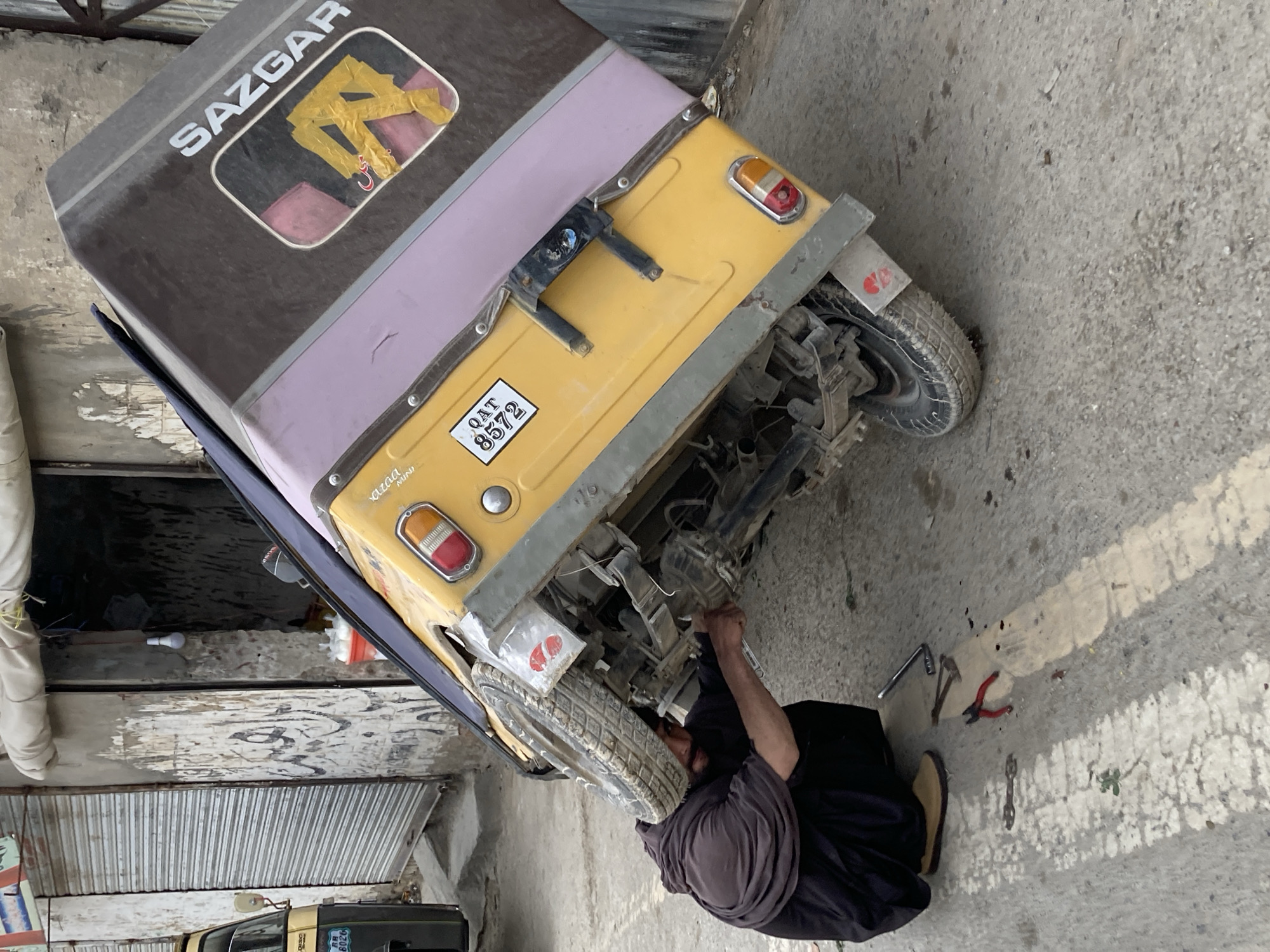 A man is repairing a Sazgar auto rickshaw on an outdoor paved surface in Quetta City Tehsil, Pakistan. The rickshaw is yellow and brown, with a license plate reading "QAT 8572". The man, wearing a dark gray shirt and a head covering, is kneeling and working on the vehicle's underside. Various tools including a wrench, pliers and hammer, are scattered on the ground near him. A building with closed shutters is in the background.