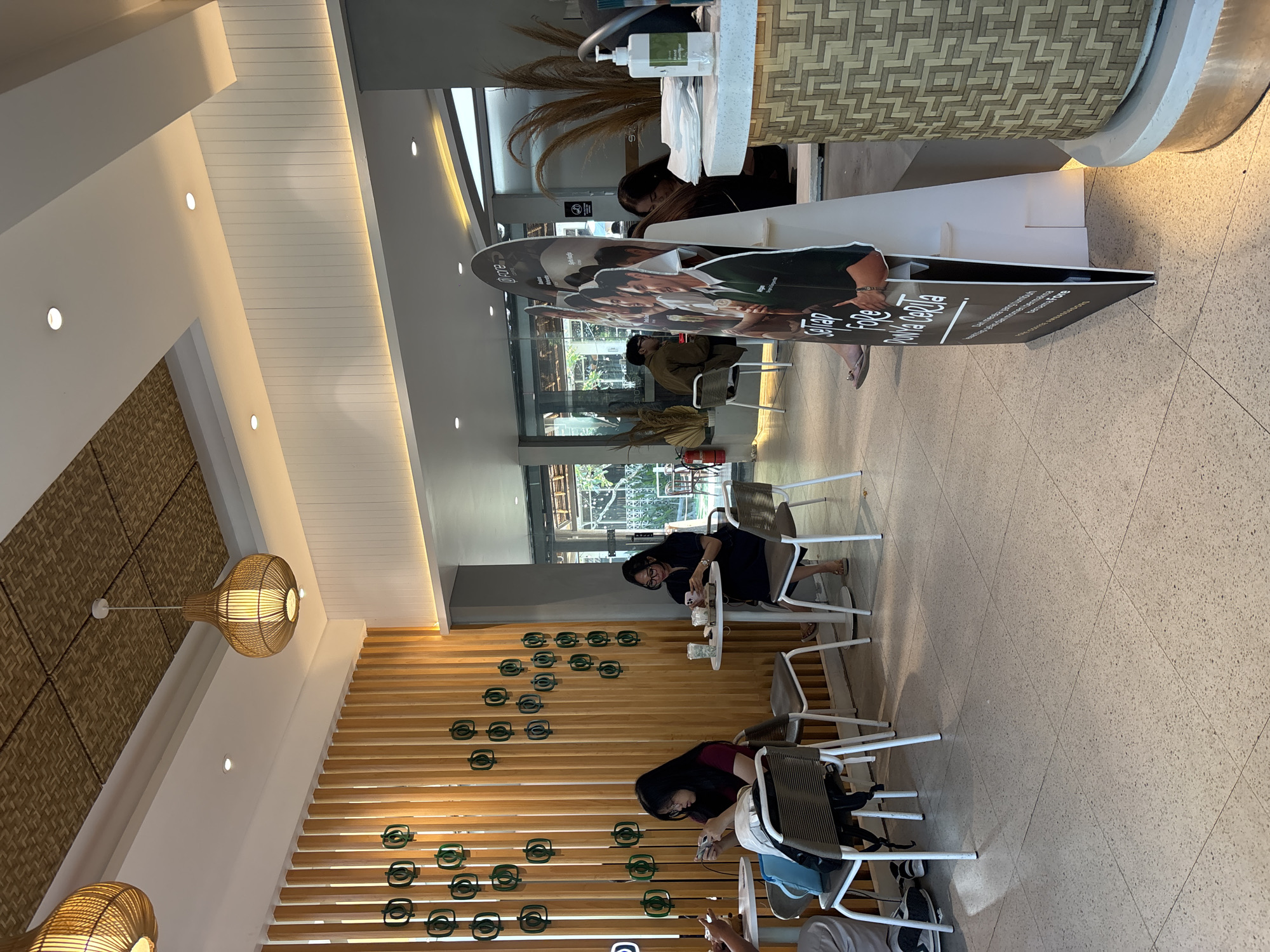The image depicts the interior of a contemporary cafe with a light and airy ambiance, likely located in Denpasar, Indonesia. The cafe features light wood paneling on the walls and ceiling, complemented by woven bamboo light fixtures that cast a warm glow. Scattered throughout the space are small, circular, green decorative elements mounted on the wood paneling.
Several patrons are seated at tables, engaged in various activities. On the left, two women are seated at a table, with one woman in a maroon shirt looking at her phone, and another in a dark top and glasses also interacting with her phone. Further back, a man is seated at a table by a window. On the right side of the image, a woman in a dark dress is seated, and a large promotional stand with images of people is prominently displayed in the foreground. A hand sanitizer dispenser is visible on a counter to the right, suggesting adherence to hygiene protocols. The flooring is a speckled terrazzo, and the overall environment suggests a relaxed and casual setting, possibly a popular spot for people to gather and socialize or work.