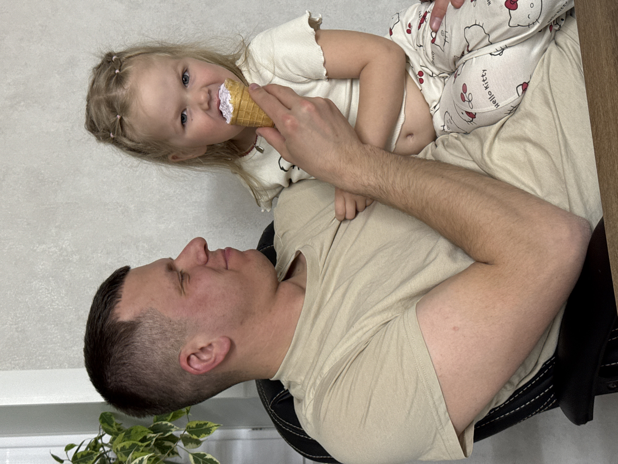 A man is sitting in a chair, and a young girl is lying on his lap. The girl is wearing Hello Kitty pajamas and a white shirt. She is eating an ice cream cone. The man is wearing a beige shirt. The background is a white wall. A plant is in the lower left corner of the image. The location of the image is Gornyi (Dvurechenskoe S/P), Russia.