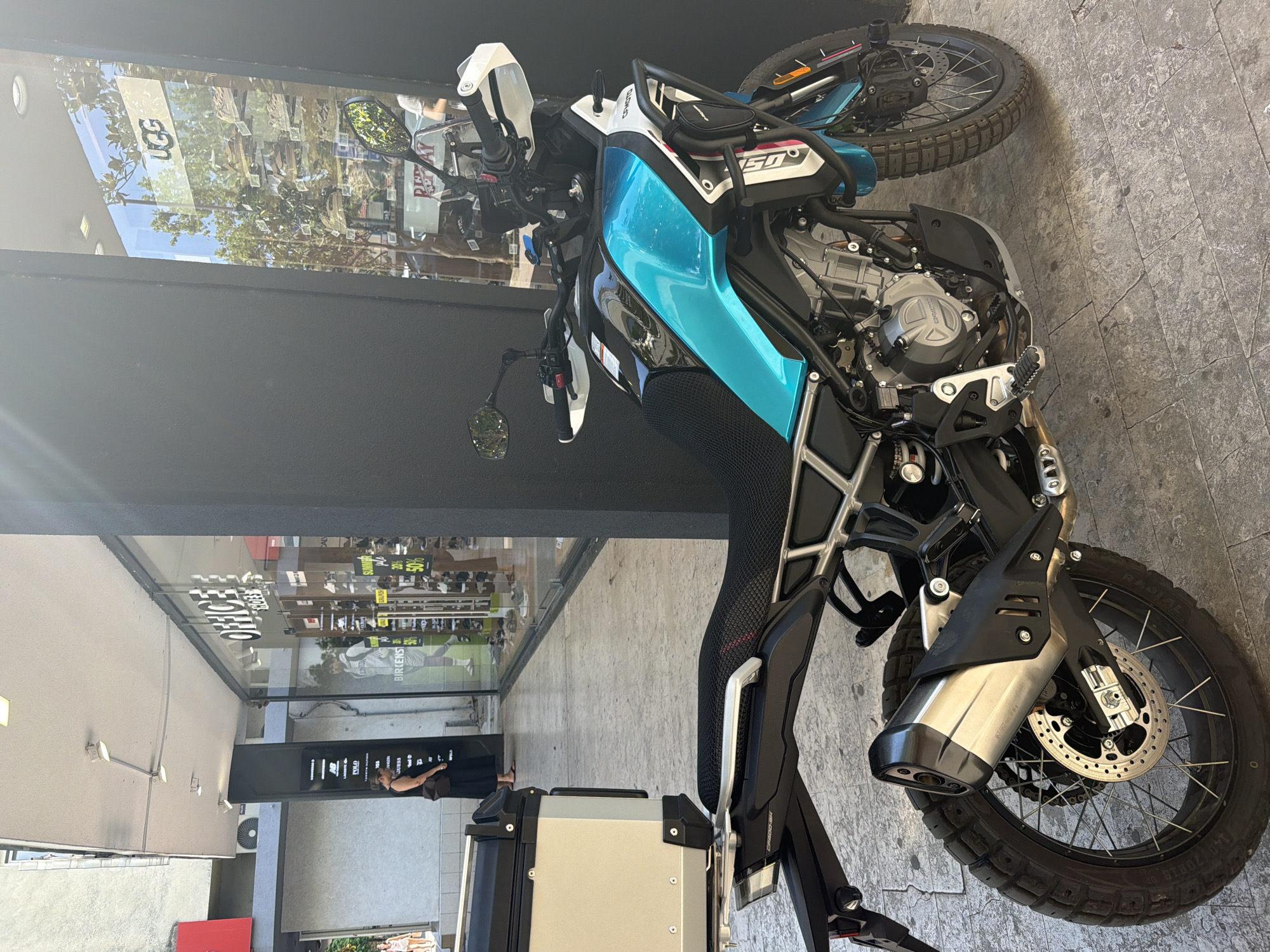 This media file shows a close-up, angled view of a blue and white adventure motorcycle parked on a grey, textured outdoor floor. The motorcycle is the primary subject. Behind it, a large storefront window reflects trees and the sky, suggesting it's daytime with good weather. The window has text visible, including "UGG" and "REPLAY". Further into the storefront, there appears to be a display of shoes or clothing. To the left of the motorcycle, a dark wall forms part of the scene. A person is partially visible in the reflection of the window, wearing dark clothing and looking towards the motorcycle. Another sign visible on the left says "OFFICE STORE". The overall impression is of a motorcycle parked outside a retail establishment on a sunny day.