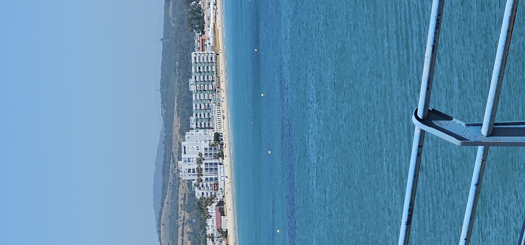 This image presents a picturesque coastal landscape, originally captured with a vertical orientation but best viewed rotated 90 degrees counter-clockwise. The scene depicts a sunny day along a beach town, consistent with the Algeciras, Spain context.
In the immediate foreground, a weathered blue metal railing, showing signs of rust and paint chipping, frames the bottom right portion of the image. This suggests the photo was taken from an elevated vantage point, such as a pier or balcony.
Beyond the railing, a wide expanse of clear, turquoise-blue ocean stretches across the midground. The water appears calm with subtle textures, and several small, bright yellow buoys are visible, floating in the water. Further towards the shore, a golden sandy beach is populated with numerous small figures of people, indicating activity like sunbathing or strolling.
Lining the beach is a strip of modern buildings, likely hotels or apartment complexes, with predominantly light-colored facades (whites, greys) and some featuring green accents. These structures create a developed urban coastline. Behind the buildings, a range of rolling hills covered in lush green vegetation rises, appearing somewhat hazy in the distance. The sky above is a uniform, clear light blue, suggesting bright, sunny weather during daytime, likely morning or early afternoon. The overall atmosphere is one of a vibrant, yet peaceful, holiday destination. No visible text is present in the image.