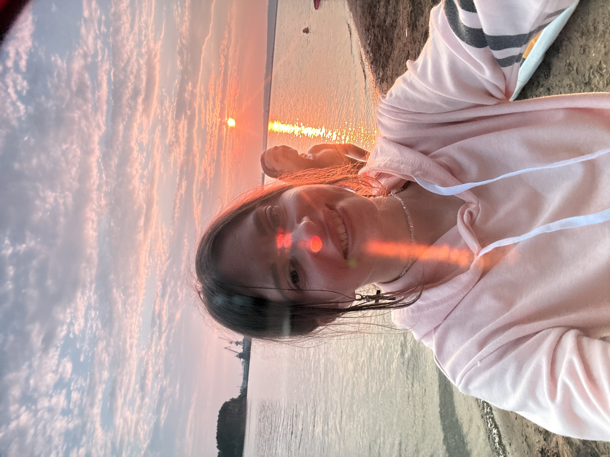 This is a portrait-oriented, low-angle selfie taken outdoors during what appears to be sunset or sunrise. The subject is a young person with long brown hair, wearing a light pink hooded sweatshirt. They are smiling directly at the camera, and their face is illuminated by the warm, golden light of the setting or rising sun, which creates a lens flare across their forehead and cheek. A delicate silver necklace is visible around their neck.
The background features a body of water, possibly a lake or the sea, with gentle ripples reflecting the sky's colors. The sky is filled with dramatic clouds, tinged with shades of pink, orange, and gold, indicative of the twilight hours. In the distance, on the left side of the frame, a silhouetted shoreline is visible with trees and industrial structures, suggesting a proximity to a port or industrial area.
The overall mood of the image is peaceful and serene, capturing a moment of personal reflection or enjoyment of the natural beauty of the sky and water. The soft lighting and the subject's happy expression contribute to a warm and inviting atmosphere. No visible text is present in the image.