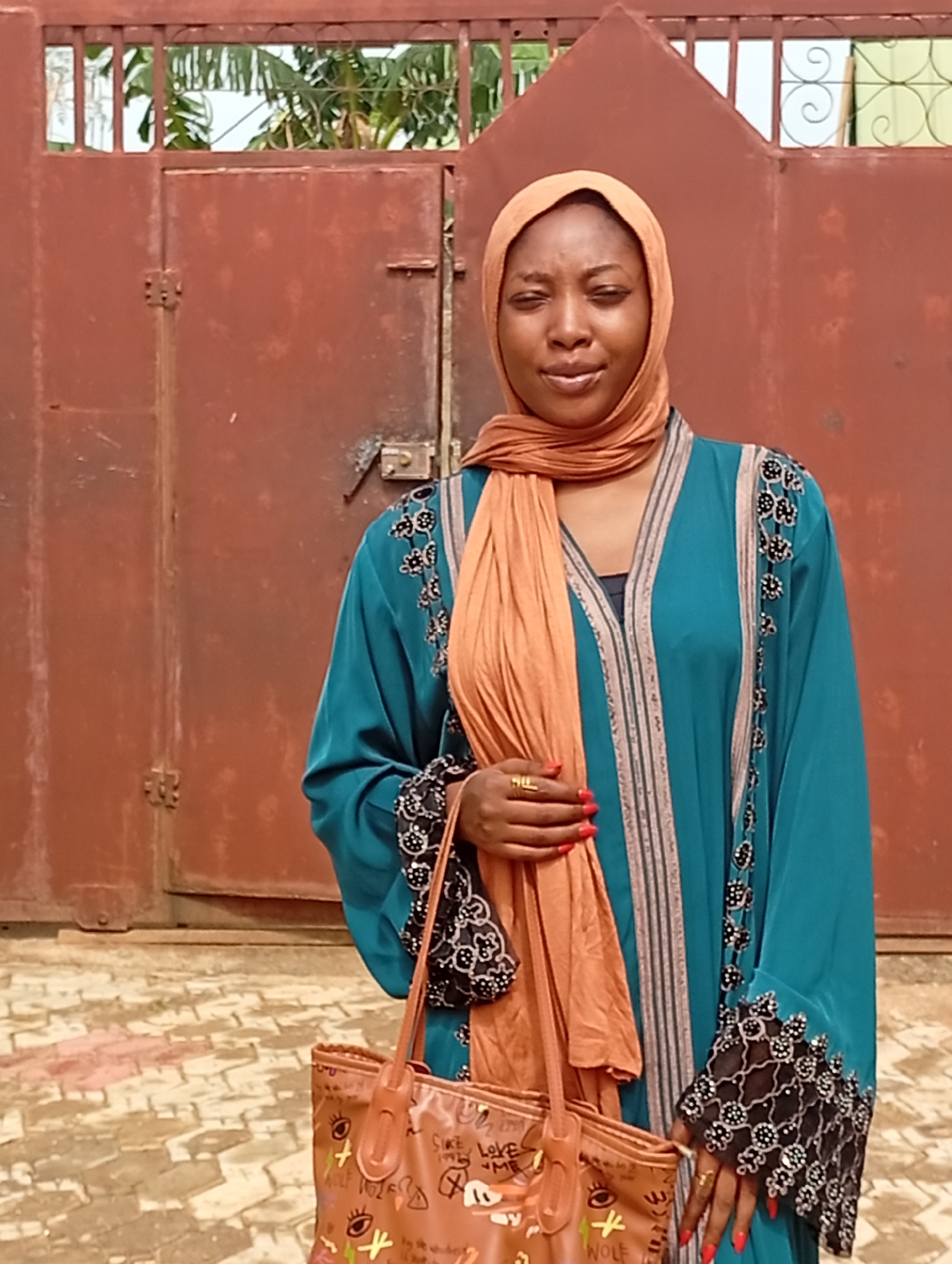 A young woman in a teal robe and beige hijab is standing in front of a rusted brown metal gate. The robe has decorative black and white patterns along the sleeves and front. She is holding a brown tote bag with illustrations. The ground is paved with reddish-brown and gray patterned bricks. The location is Unknown, Unknown.