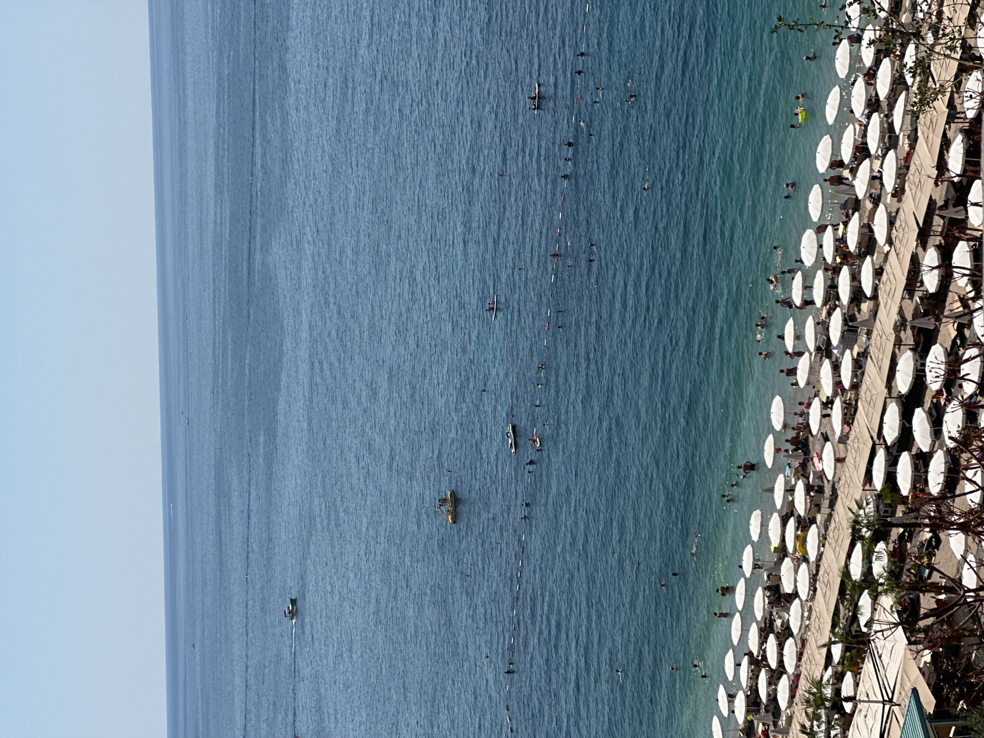 The image depicts a crowded beach scene in Kamenovo, Montenegro, on a clear, sunny day. The foreground is dominated by rows of white beach umbrellas and lounge chairs, some occupied by beachgoers. Beyond the beach, the clear blue Adriatic Sea is visible, dotted with several boats and numerous swimmers and people engaged in water activities. A buoys marking a swimming area are also visible in the water. The horizon is clear, indicating good weather conditions. The scene is bustling with activity, capturing the essence of a lively beach resort.