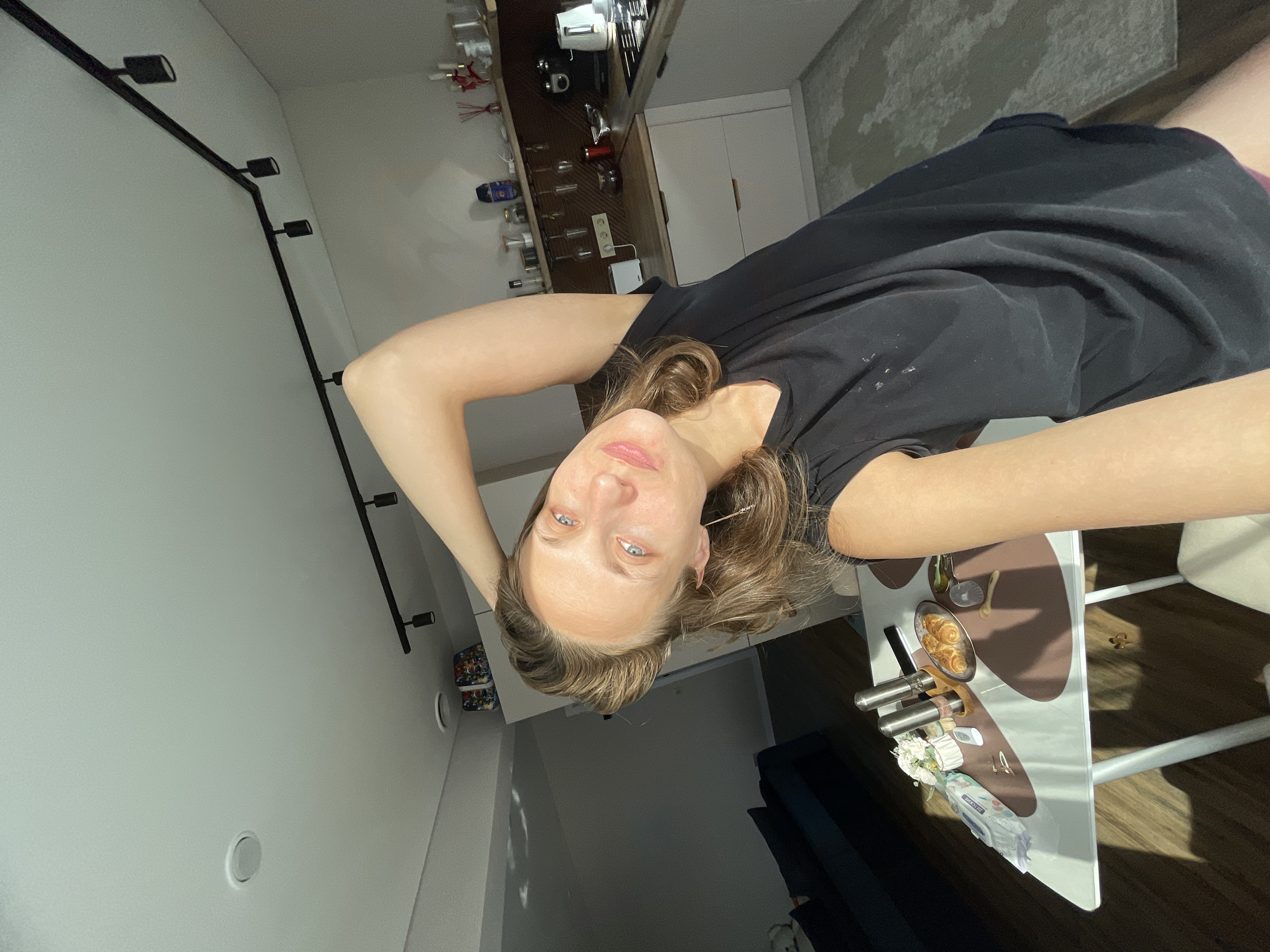 A woman is taking a selfie in a kitchen. She has long brown hair and blue eyes and is wearing a black t-shirt. Her arm is raised behind her head. On a table in front of her, there are croissants, a wet wipe package, salt and pepper shakers, and a small vase of flowers. The kitchen has white cabinets and a dark countertop. A window casts shadows across the floor. There is a dark blue couch in the background. The scene is located in the city of Podshivalovo, Russia.