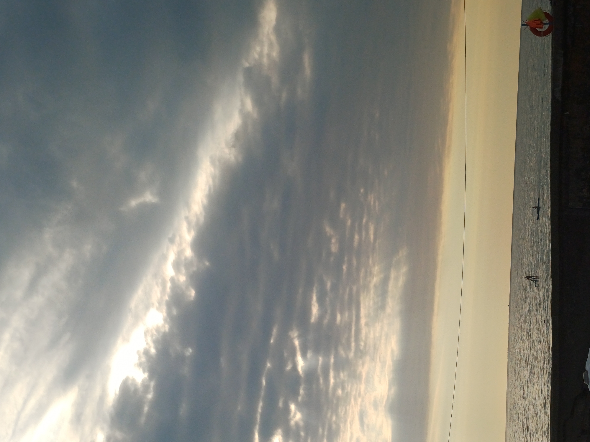 The image depicts a view of the Black Sea from a waterfront promenade in Sochi, Russia. The sky is filled with dramatic, dark clouds, with shafts of sunlight breaking through in the upper left portion of the frame, creating a textured, layered effect. The sea is calm, with small waves rippling the surface. In the mid-ground, two individuals are visible on paddleboards. One paddleboarder is standing, while the other is seated. A life preserver, yellow and orange, hangs on the right side of the frame near the edge of the promenade. The promenade itself is constructed of dark stone. The horizon is a clear line separating the water and the sky. The overall atmosphere is one of impending weather change, with the sunlight suggesting a break in the clouds.