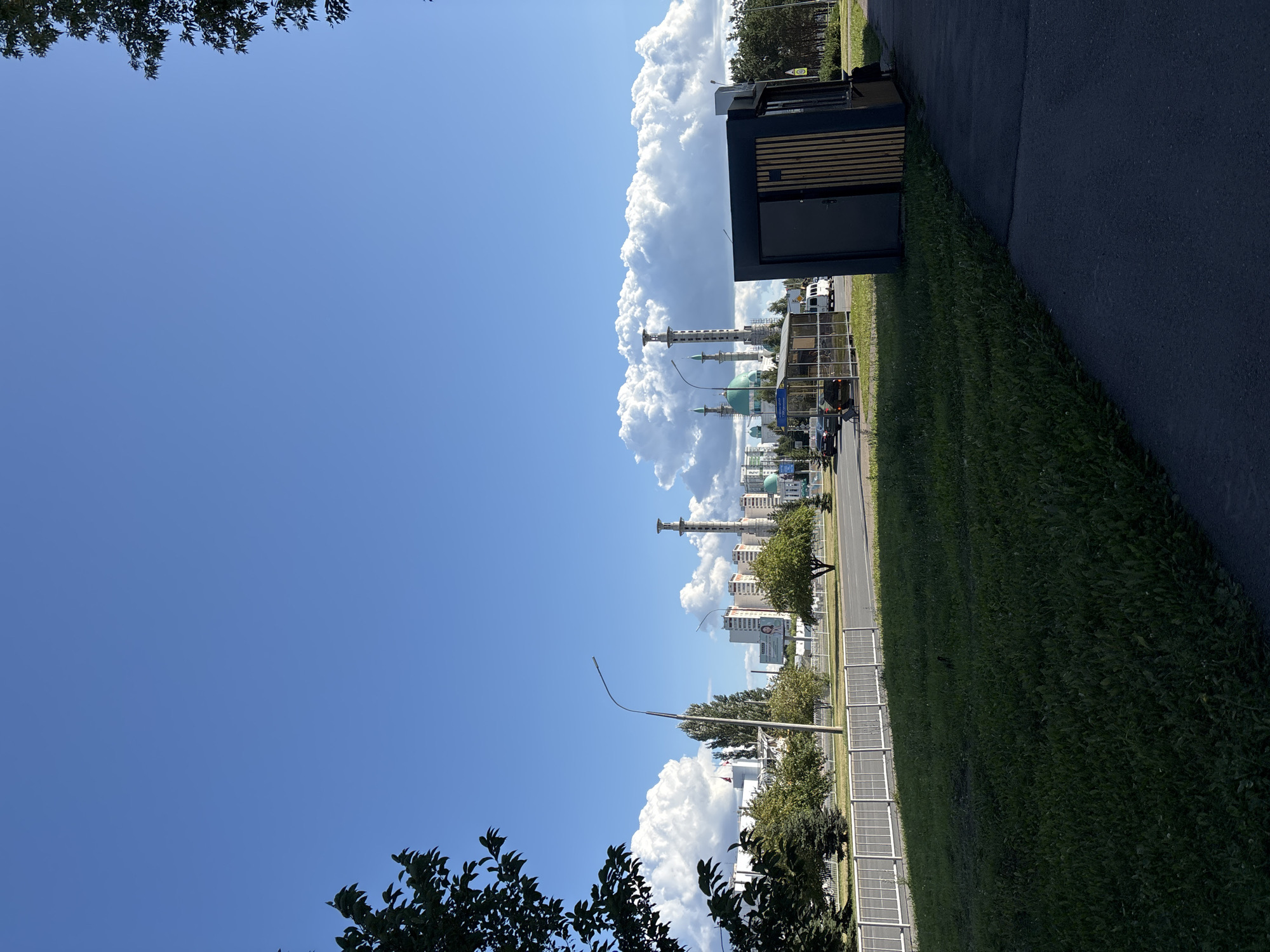 The image depicts an outdoor scene with a clear blue sky dotted with fluffy white clouds. In the foreground, a paved sidewalk runs diagonally from the bottom right corner. Adjacent to the sidewalk is a grassy area. To the left of the sidewalk is a small, modern-looking kiosk with dark siding and wooden accents, featuring a closed door and a small window. Beyond the kiosk, a road is visible with a few cars and a bus stop shelter. Across the road, a white metal fence separates the road from a green space with trees and a few buildings in the background. Further in the distance, a complex of buildings with green domes and tall, slender towers, characteristic of a mosque under construction, is prominent. The city of Naberezhnye Chelny, Russia, is identifiable by the distinctive architecture of the mosque complex. The overall scene suggests a sunny day in an urban environment.