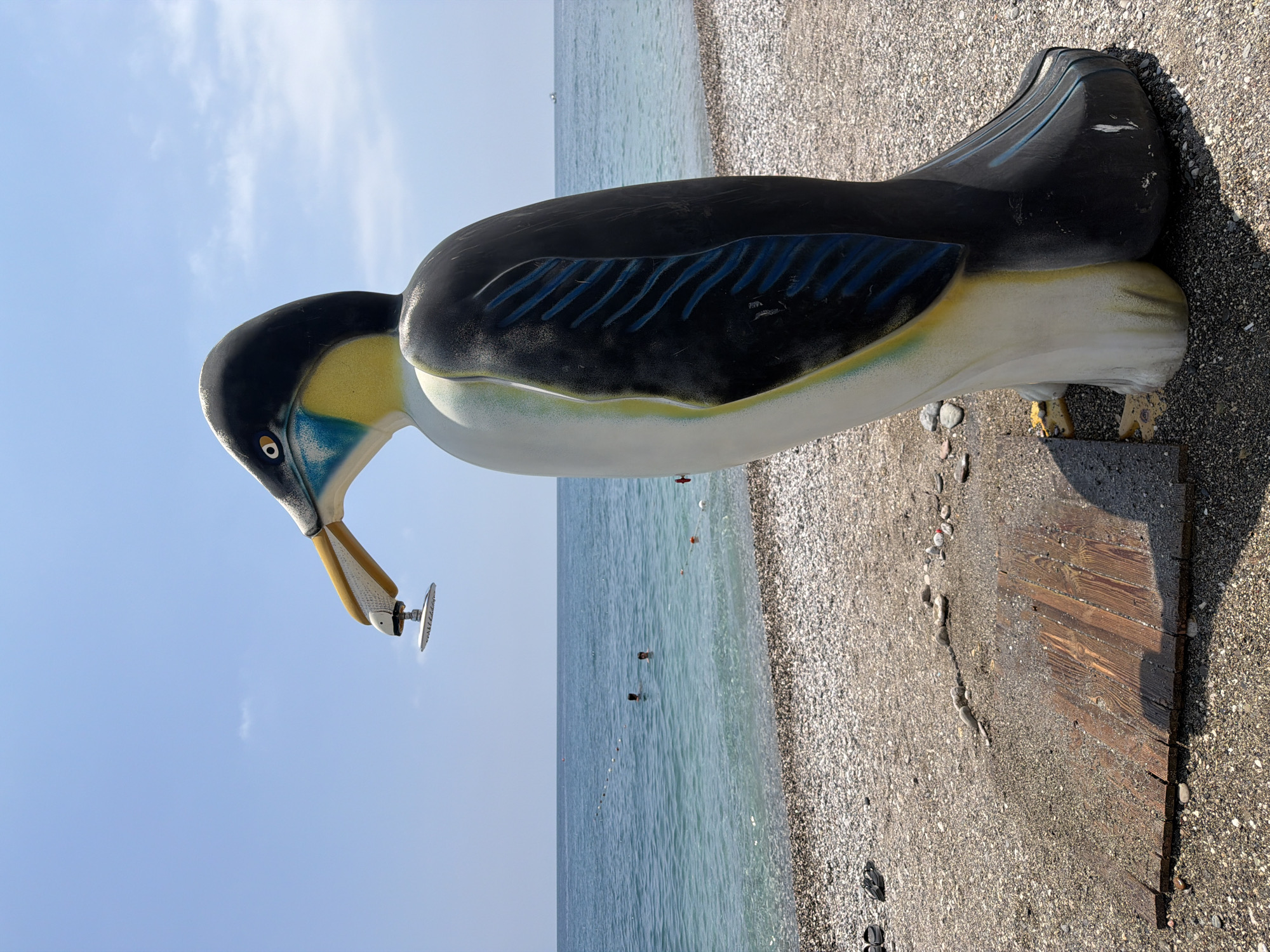 A large, colorful statue of a penguin stands on a pebbled beach in Kemer, Turkey. The penguin statue is predominantly white and black, with blue and yellow accents on its face and wings. It is positioned to the right of the frame, facing left, with its beak open. A shower head is attached to the beak of the penguin, suggesting it may function as an outdoor shower.
In the background, the turquoise water of the sea stretches to the horizon under a clear blue sky. Two individuals are visible swimming in the ocean. The beach is covered in small, smooth pebbles, and a wooden plank is laid on the ground in front of the penguin statue. A few scattered pebbles and what appear to be discarded plastic bottles are also visible on the beach. The lighting suggests it is daytime, with a slight shadow cast by the penguin statue onto the wooden plank.