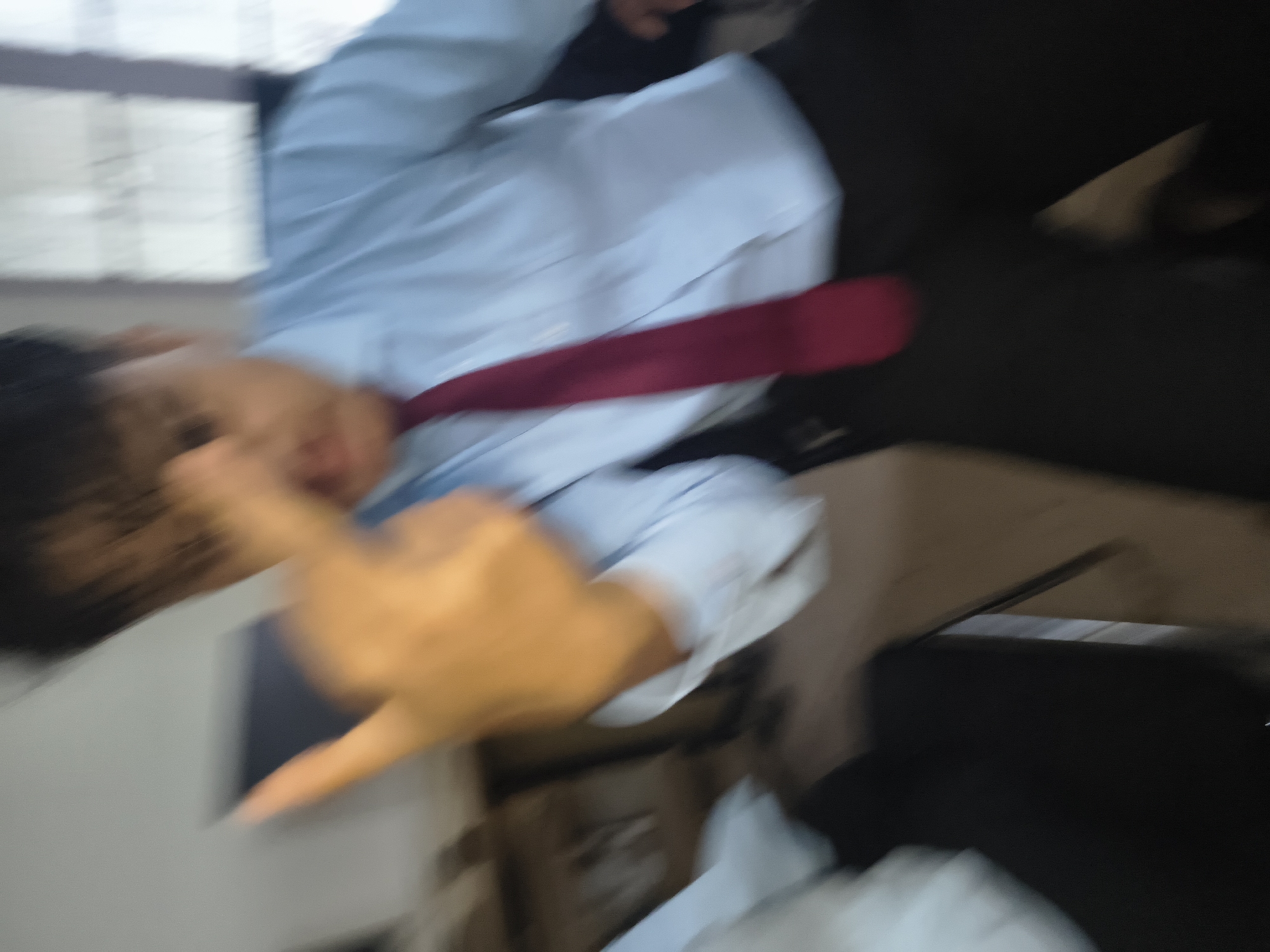 The image shows a person in a light blue dress shirt and a red tie, blurred due to motion. The person's right hand is raised, with the middle finger extended, in a gesture. The background is out of focus, but appears to be an indoor setting with light-colored walls and possibly furniture. There are no specific landmarks or visual cues that can be used to determine the location of Dharamshala, India.
