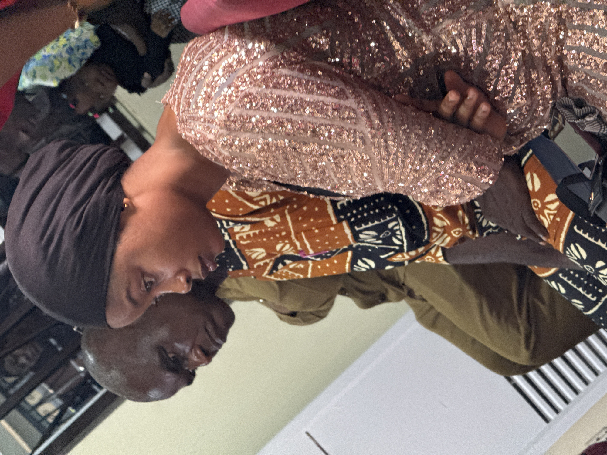 The image shows two individuals in an indoor setting, potentially within the city of Lugbe, Nigeria, though this cannot be definitively confirmed. The primary subject is a woman wearing a dark head covering and an outfit with an ornate sequined top and a patterned textile underneath. A second person, a bald male, is positioned adjacent to the woman. The background is generic and does not reveal specific details.