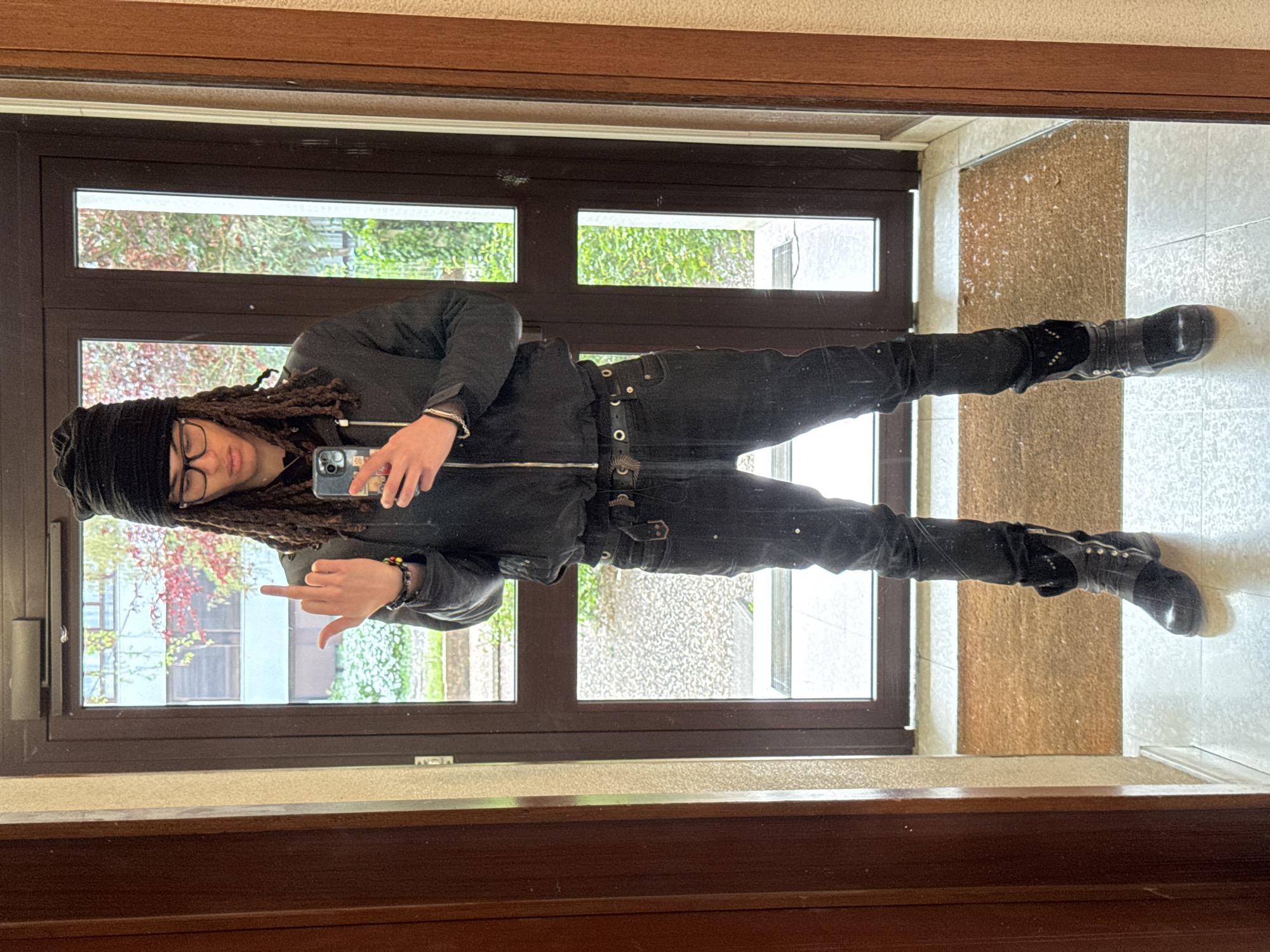 A person with dreadlocks and a black beanie is taking a mirror selfie while giving the middle finger. They are wearing glasses, a black jacket, black pants with a studded belt, and black boots. The reflection in the mirror shows a building with a window and greenery outside. The location appears to be inside a building in Les Ulis, France, based on the window view. The background also shows tiled walls and a door frame.